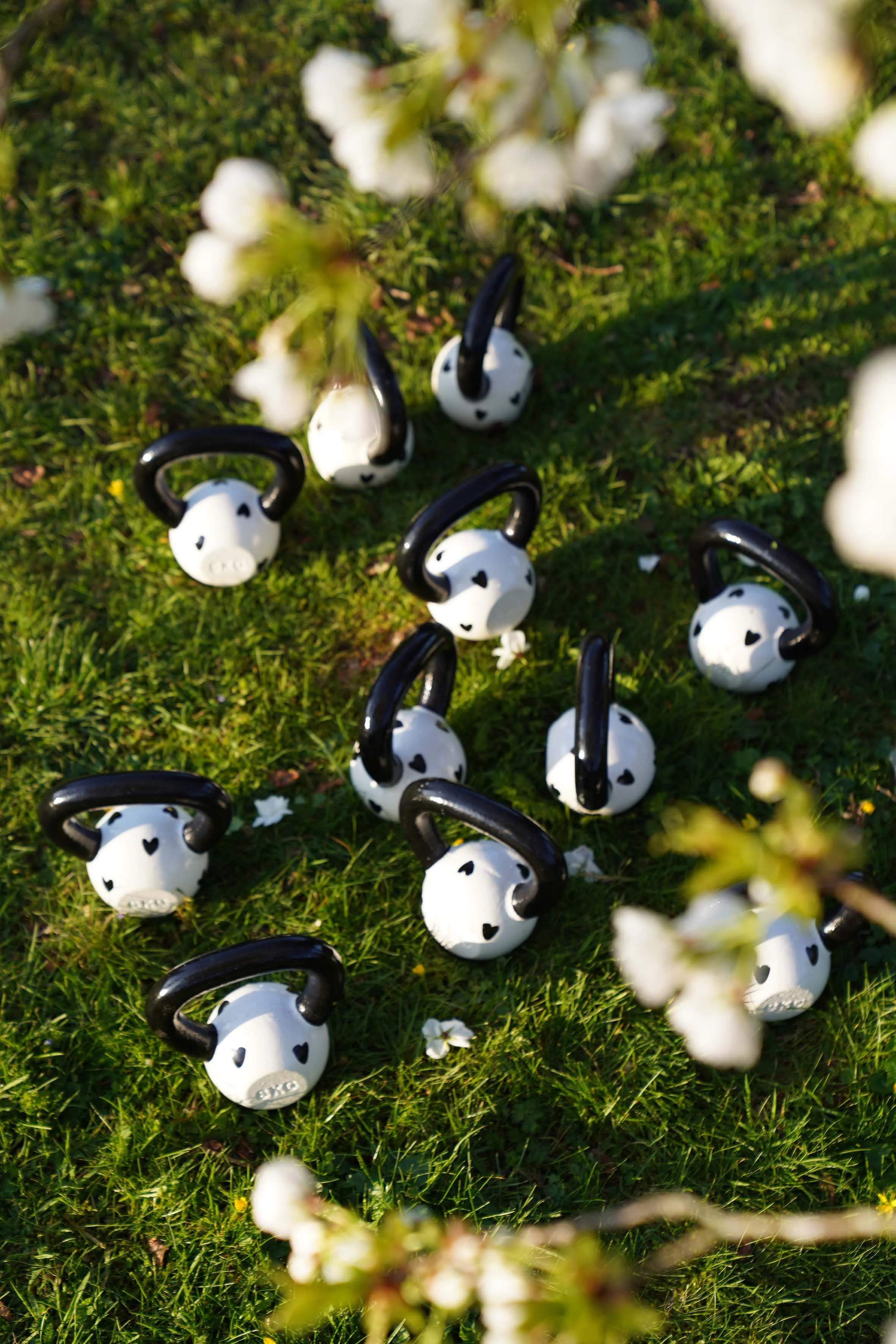 Several white kettlebell weights with black handles on green grass, partially obscured by blurred white flowers in the foreground.