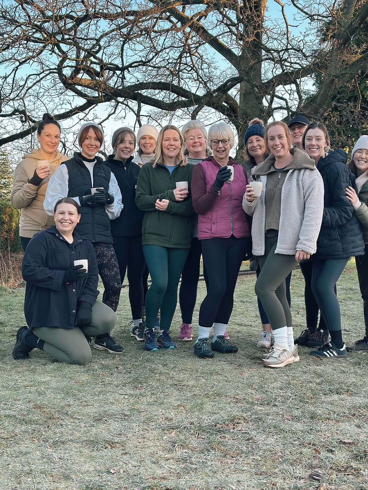 A group of women standing outdoors on grass, holding cups, dressed in casual athletic clothing, smiling, in front of a large tree with bare branches.