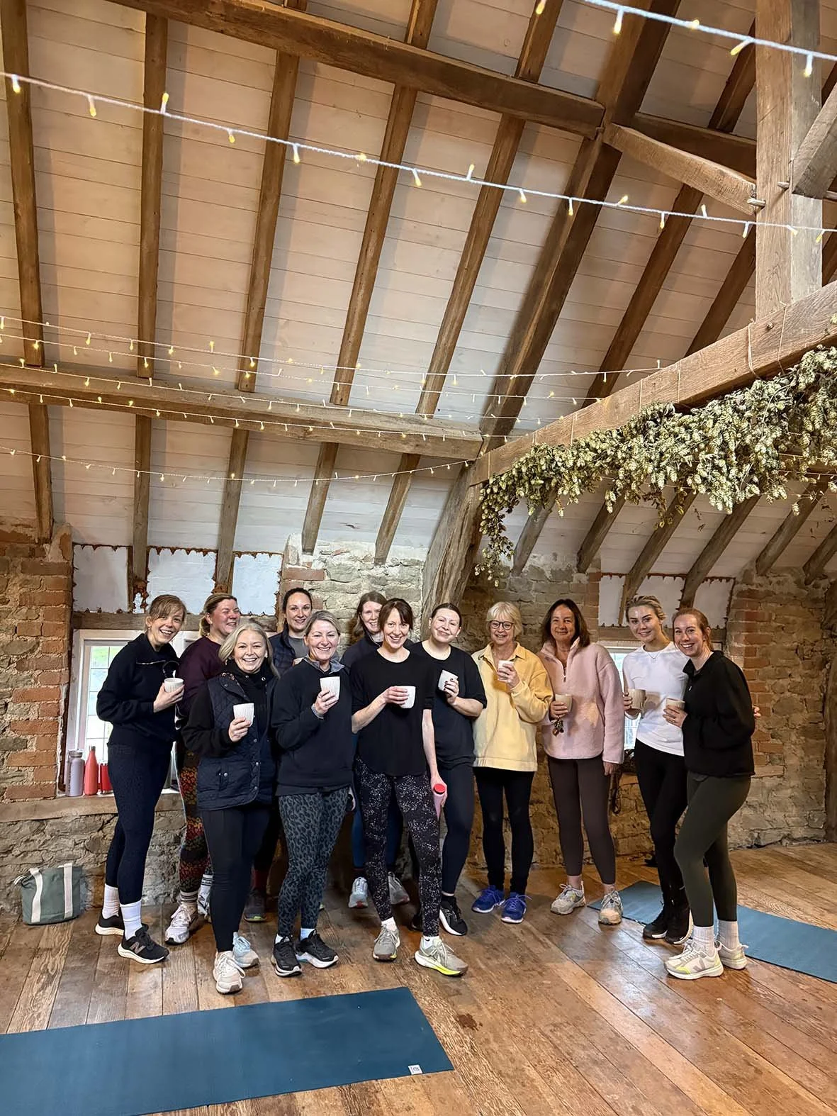 A group of women in casual clothing standing together in a rustic room with brick walls and wooden beams, holding drinks and smiling.
