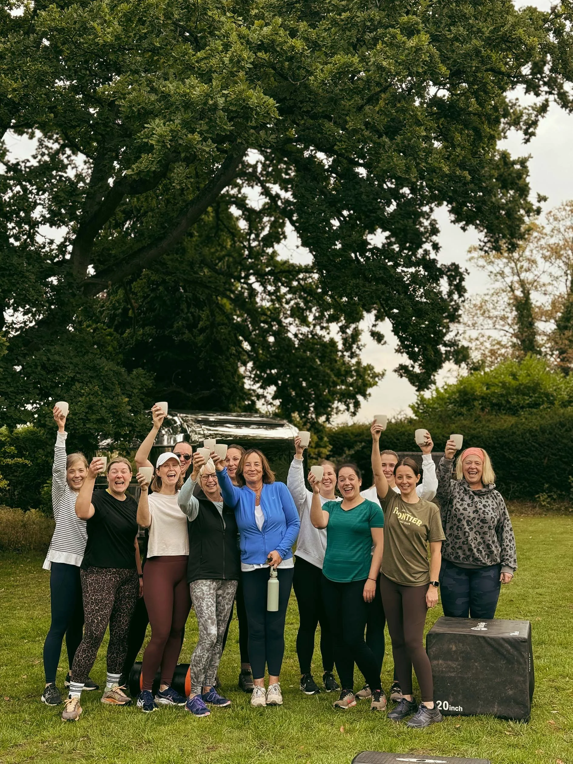 A group of diverse women outdoors holding up teacups, smiling, in a park with grass, trees, and cloudy sky.