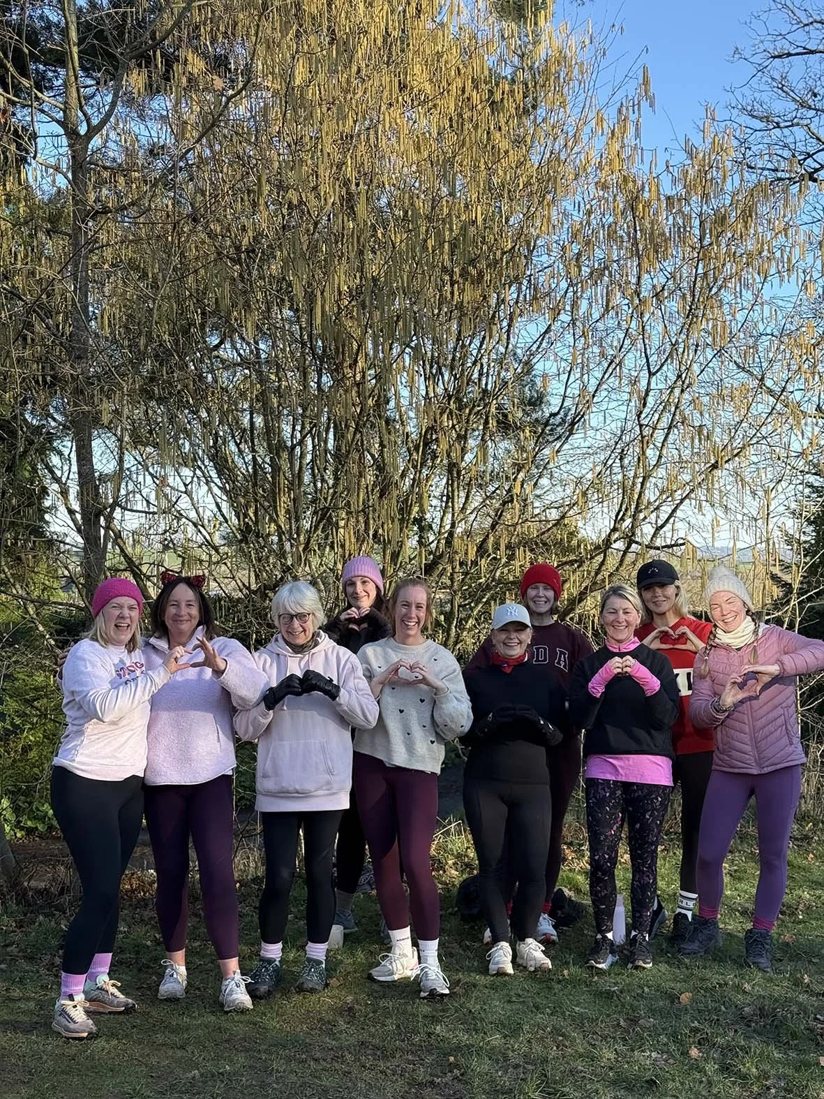 Group of nine women standing outdoors in front of a large tree, smiling and making heart shapes with their hands, dressed in workout clothes and colorful hats.