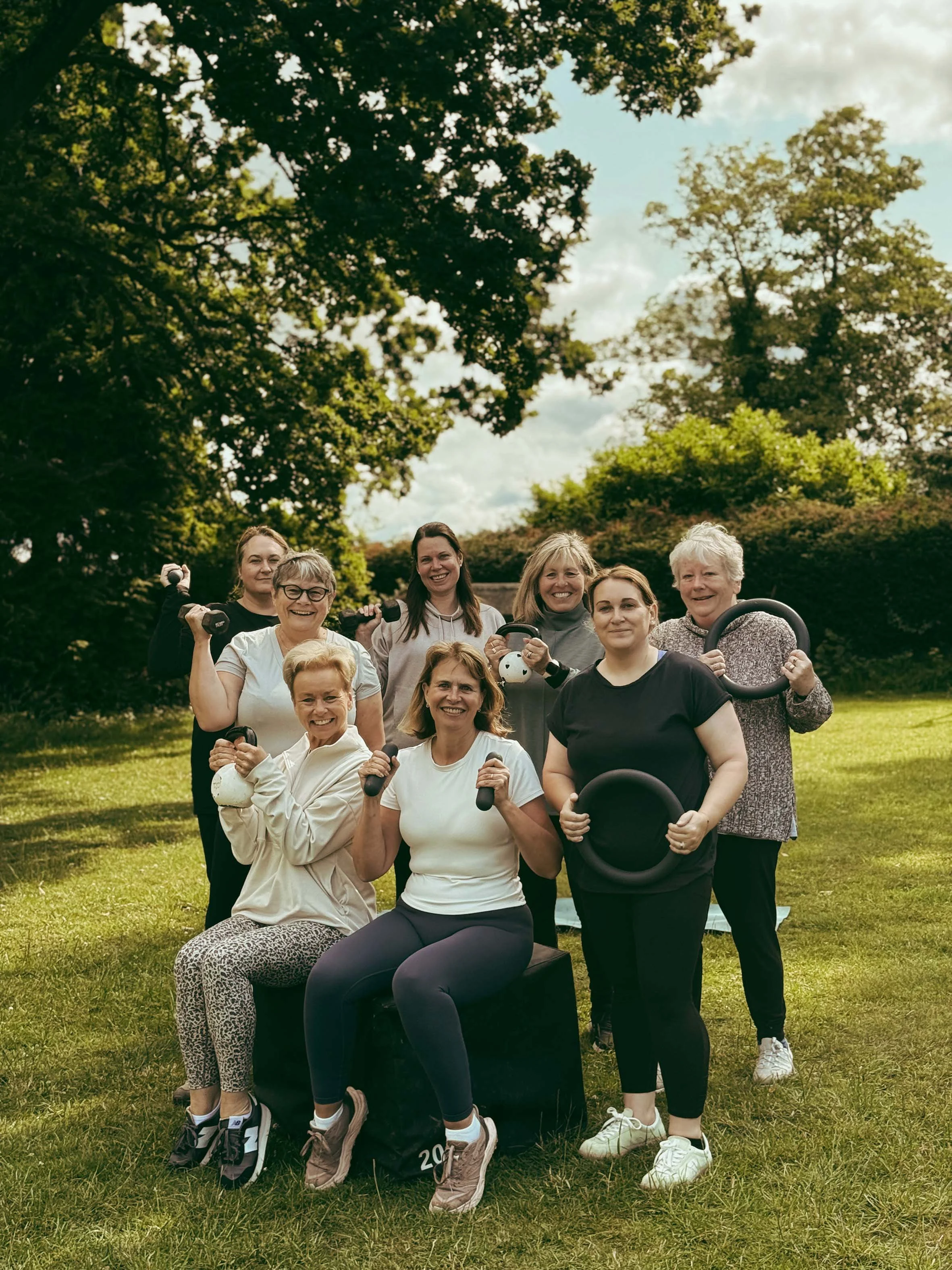 Group of smiling women holding fitness equipment outdoors in a park with green trees and grass.
