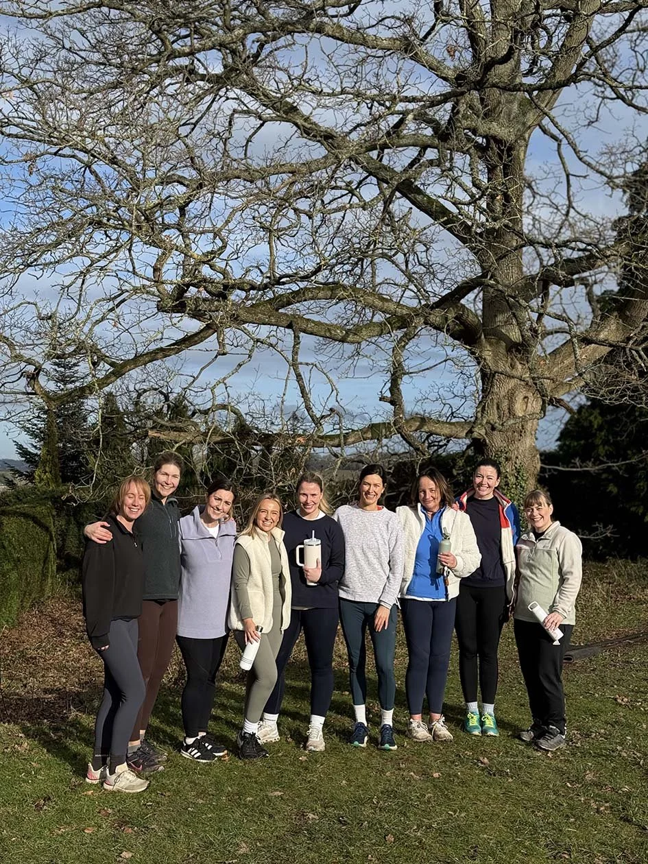 Eight women standing outdoors in athletic clothing, smiling, with a large leafless tree in the background.