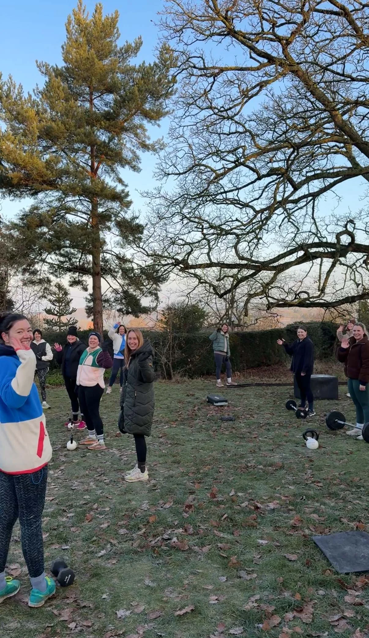 A group of people outdoors in a park, smiling and waving, with some fitness equipment like dumbbells and kettlebells on the grass, trees in the background, and a clear sky overhead.