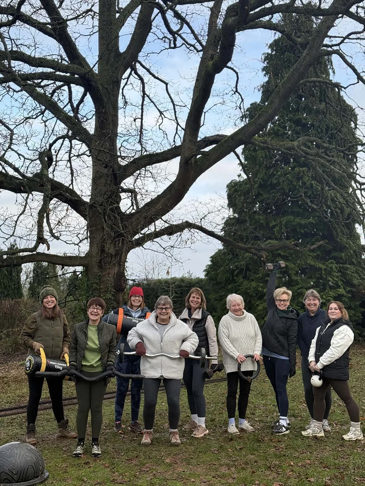 Group of women outdoors in front of a large leafless tree, smiling and posing for the photo. Some are holding workout equipment, and one woman is raising her fist in celebration.