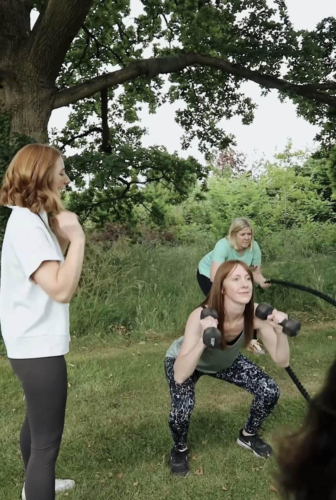 Four women exercising outdoors under a large tree. One woman is holding dumbbells, another is kneeled holding a rope, a third is standing and watching, and the fourth is crouching with a jump rope.