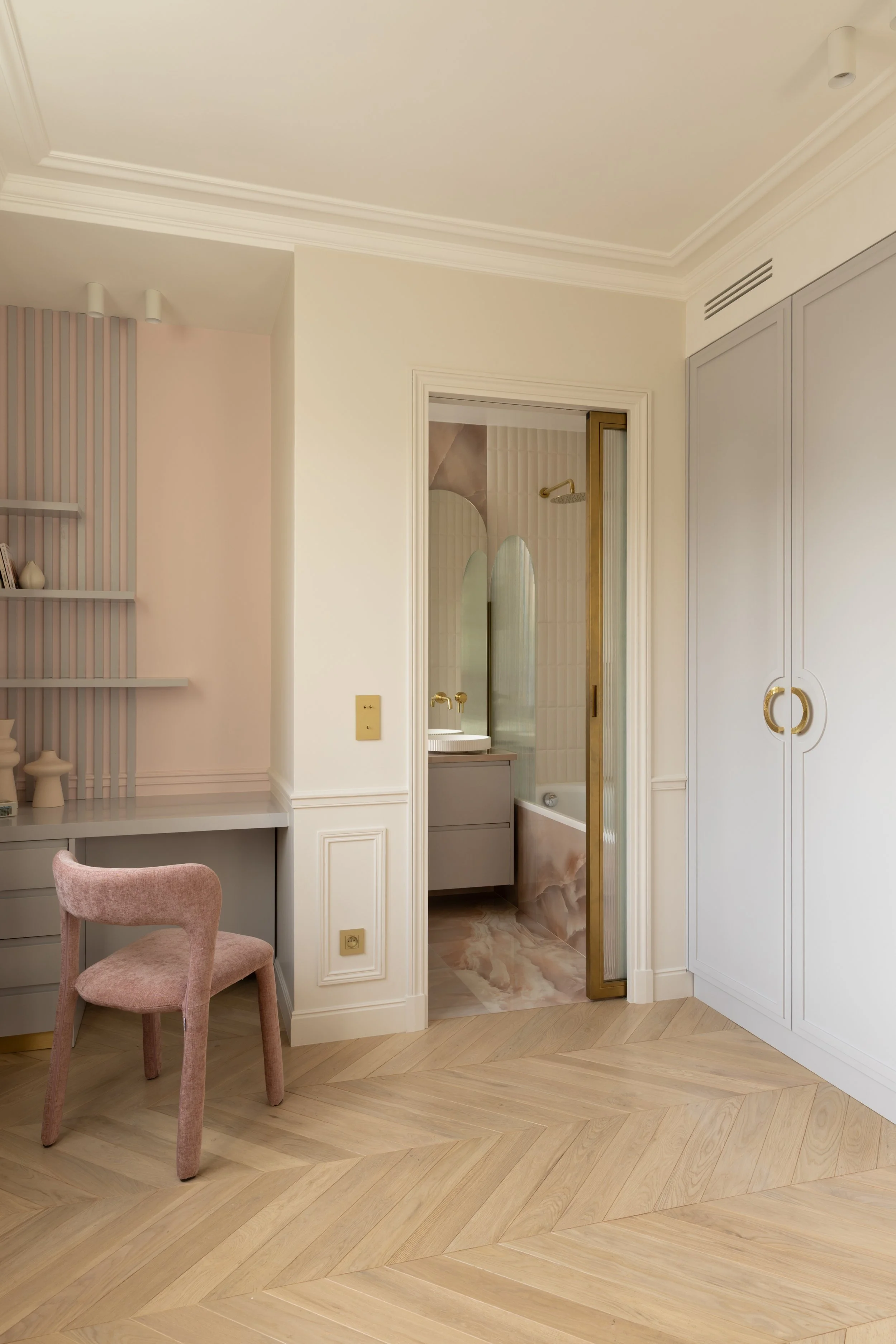 Chambre enfant élégante d’un appartement contemporain avec parquet en chevrons, mobilier sur mesure gris souris et chaise en velours rose poudré. Une porte à galandage en verre flute ouverte sur une salle de bain design en Onix rose, avec miroir arch