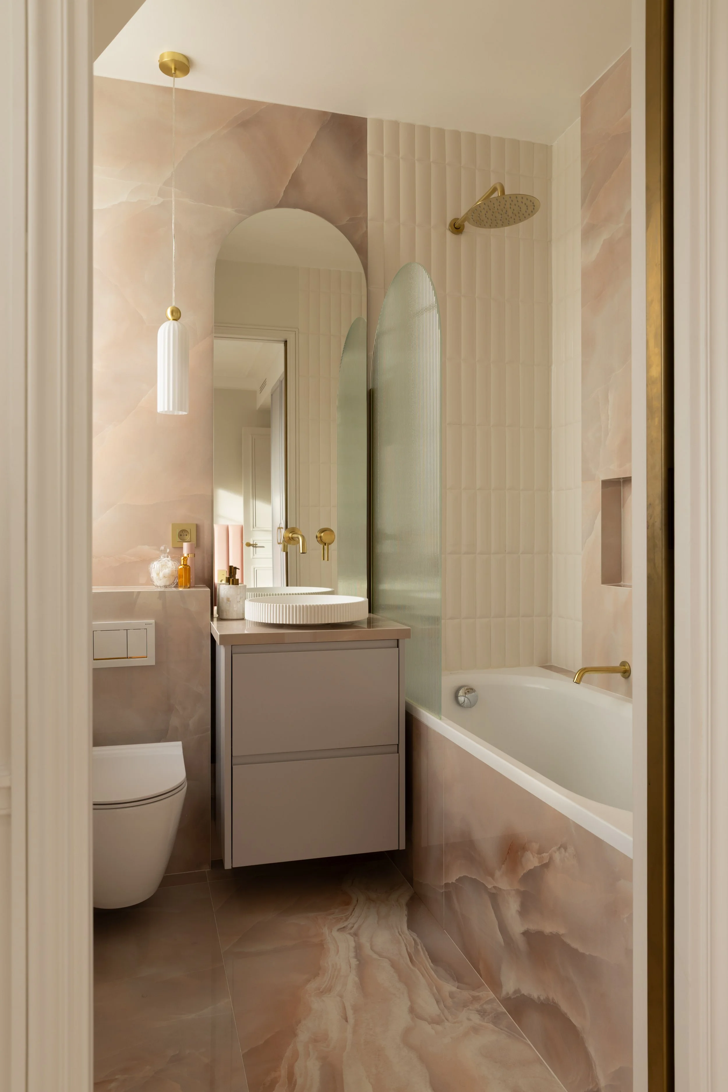 Salle de bain moderne et raffinée avec baignoire encastrée, meuble vasque suspendu gris souris et miroir vertical rétroéclairé. Les murs en pierre naturelle aux tons beige rosé et les lignes architecturales apportent une atmosphère luxueuse et contem