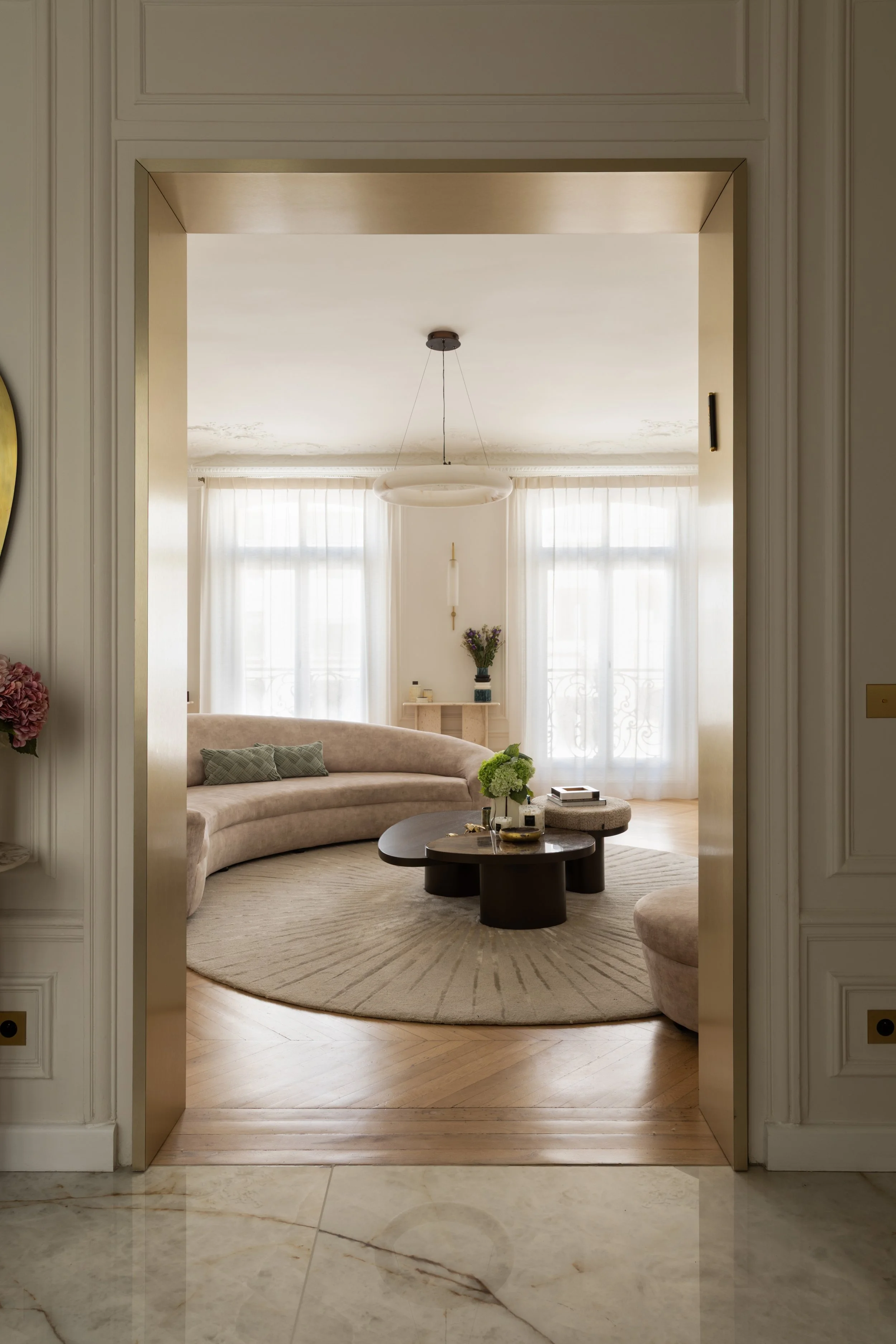 Vue depuis l’entrée vers le salon du projet Malesherbes à Paris : encadrement doré, canapé arrondi beige, tables basses sculpturales et parquet en point de Hongrie — Intérieurs Chomet.