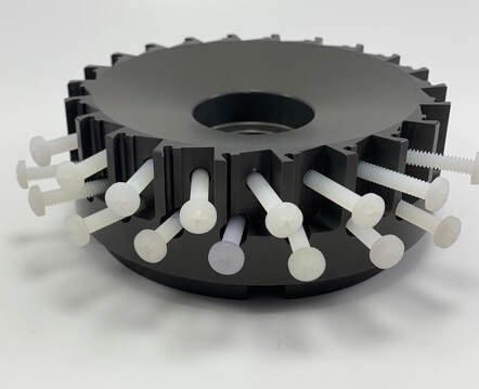 A black circular object with multiple white plastic pegs extending outward from the edge.