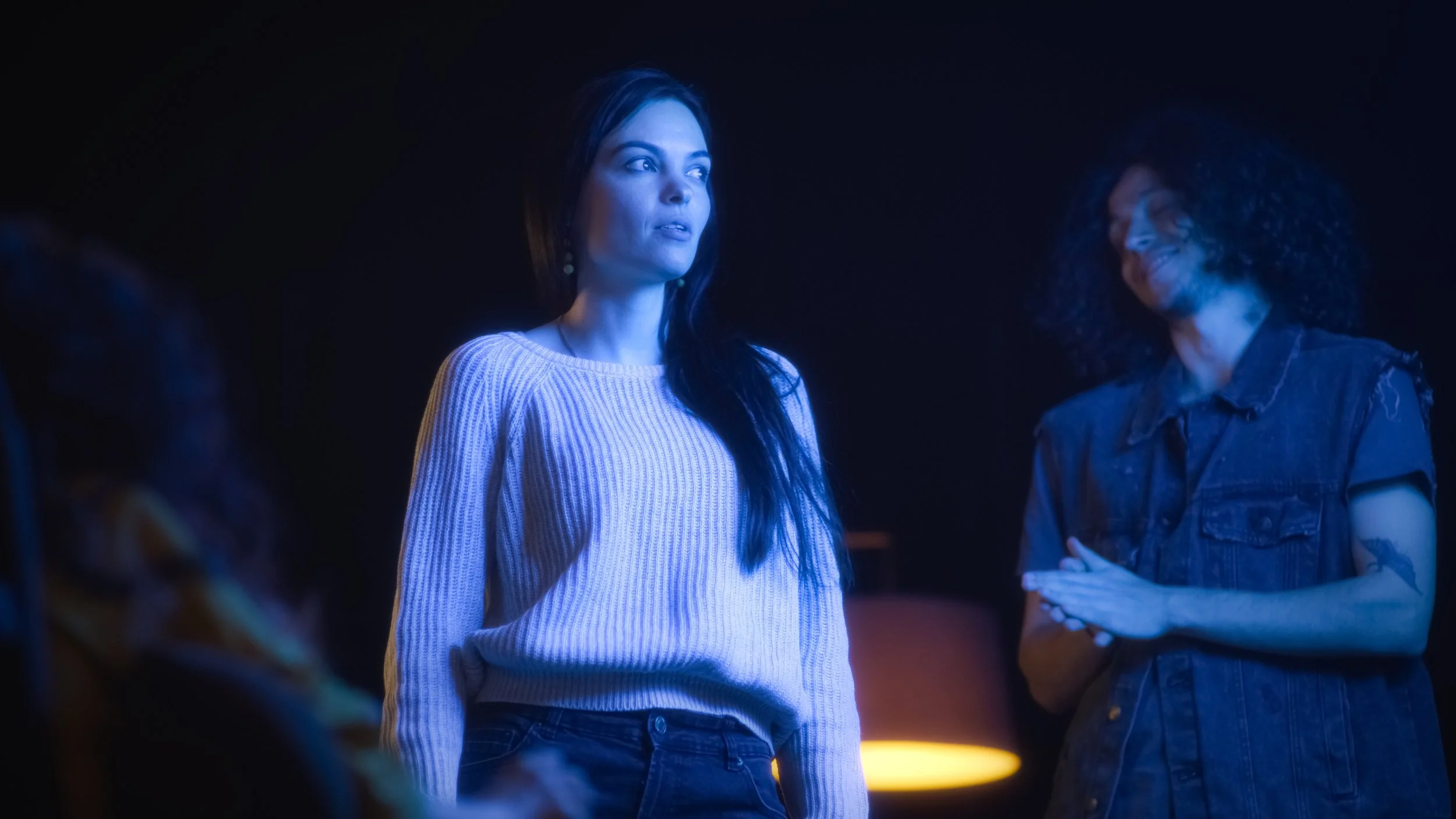 A woman in a white sweater with long dark hair is looking to the side, illuminated by blue lighting. She is standing with another woman who has curly hair and is wearing a denim jacket, also smiling. The background is dark with a hint of orange light.