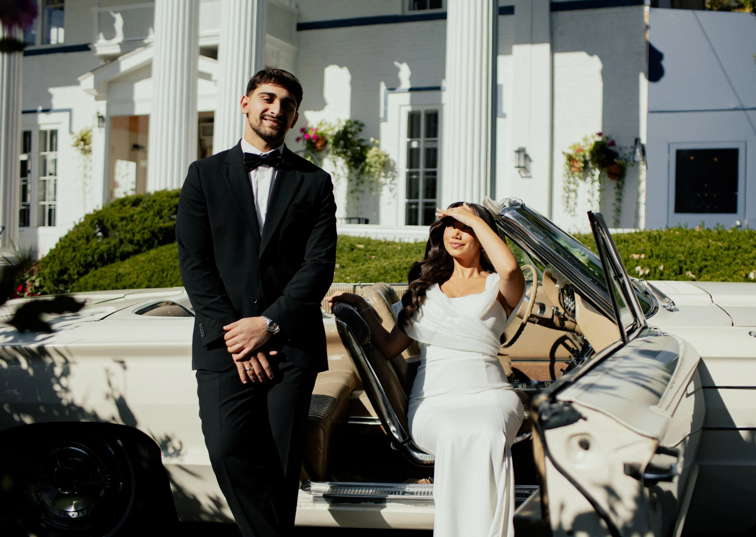 A wedding couple in front of a white mansion. The groom in a black tuxedo with a bowtie is standing, smiling, while the bride in a white dress is sitting in a vintage convertible car, shielding her eyes from the sun with her hand.