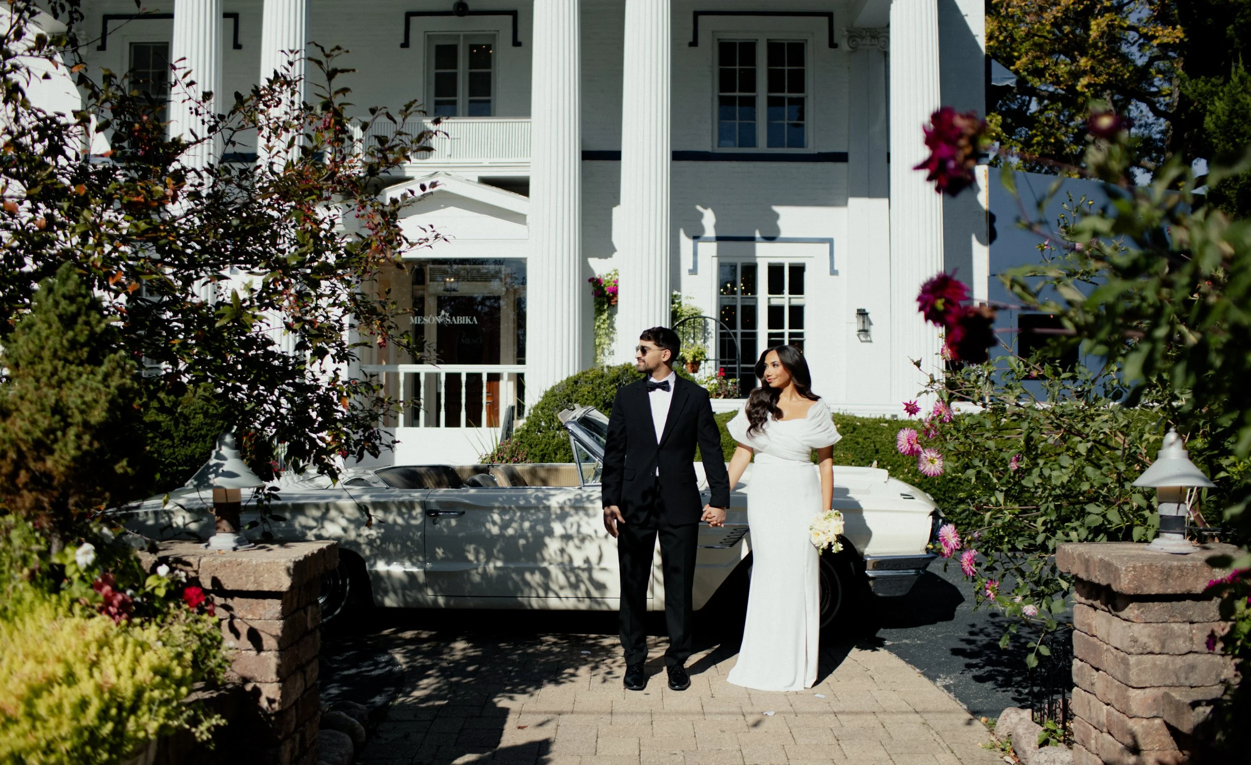 A bride and groom holding hands in front of a vintage white convertible car, outside a large white mansion with pillars, surrounded by greenery and blooming flowers.