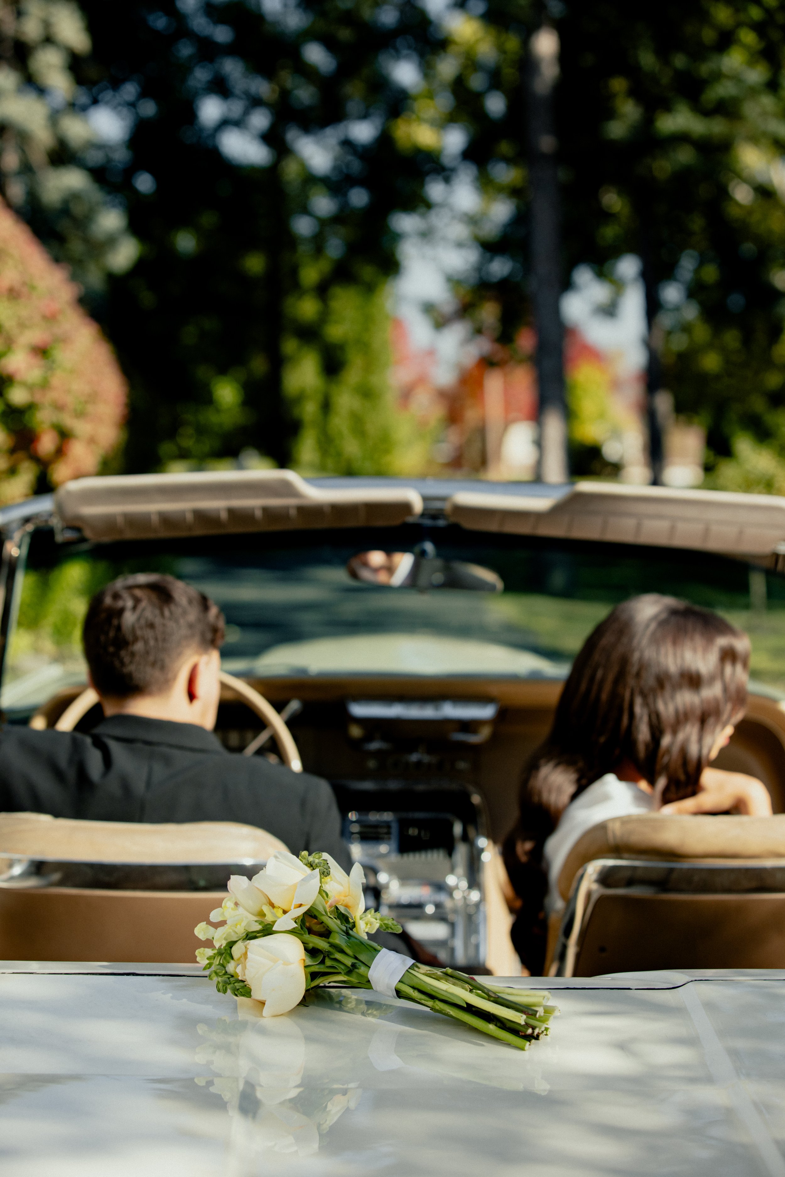 A bouquet of white calla lilies on the trunk of a car, with a couple sitting inside a vintage convertible car in a park-like setting.