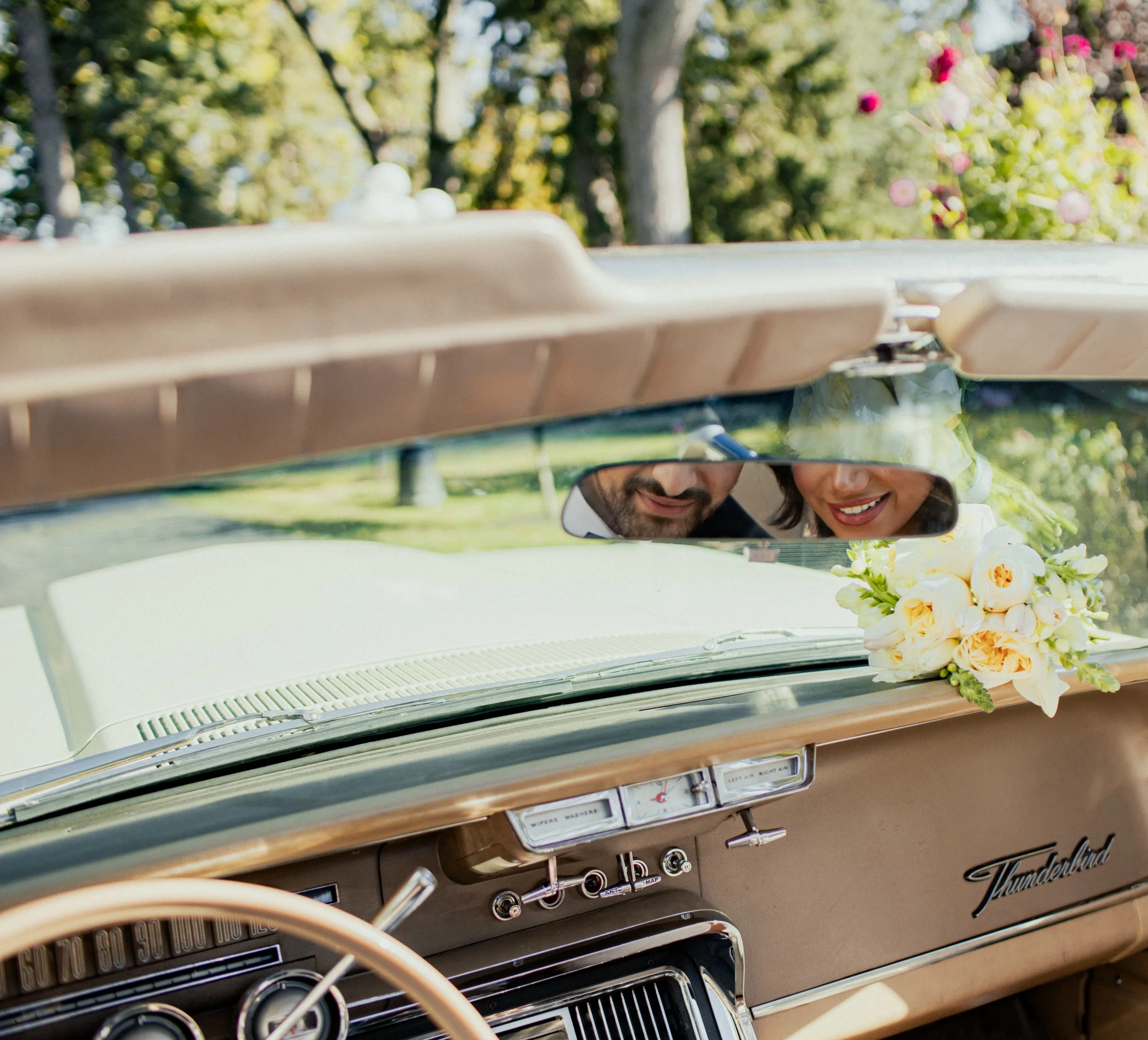 A couple peering over the dashboard of a vintage car decorated with flowers, with trees and flowers in the background.