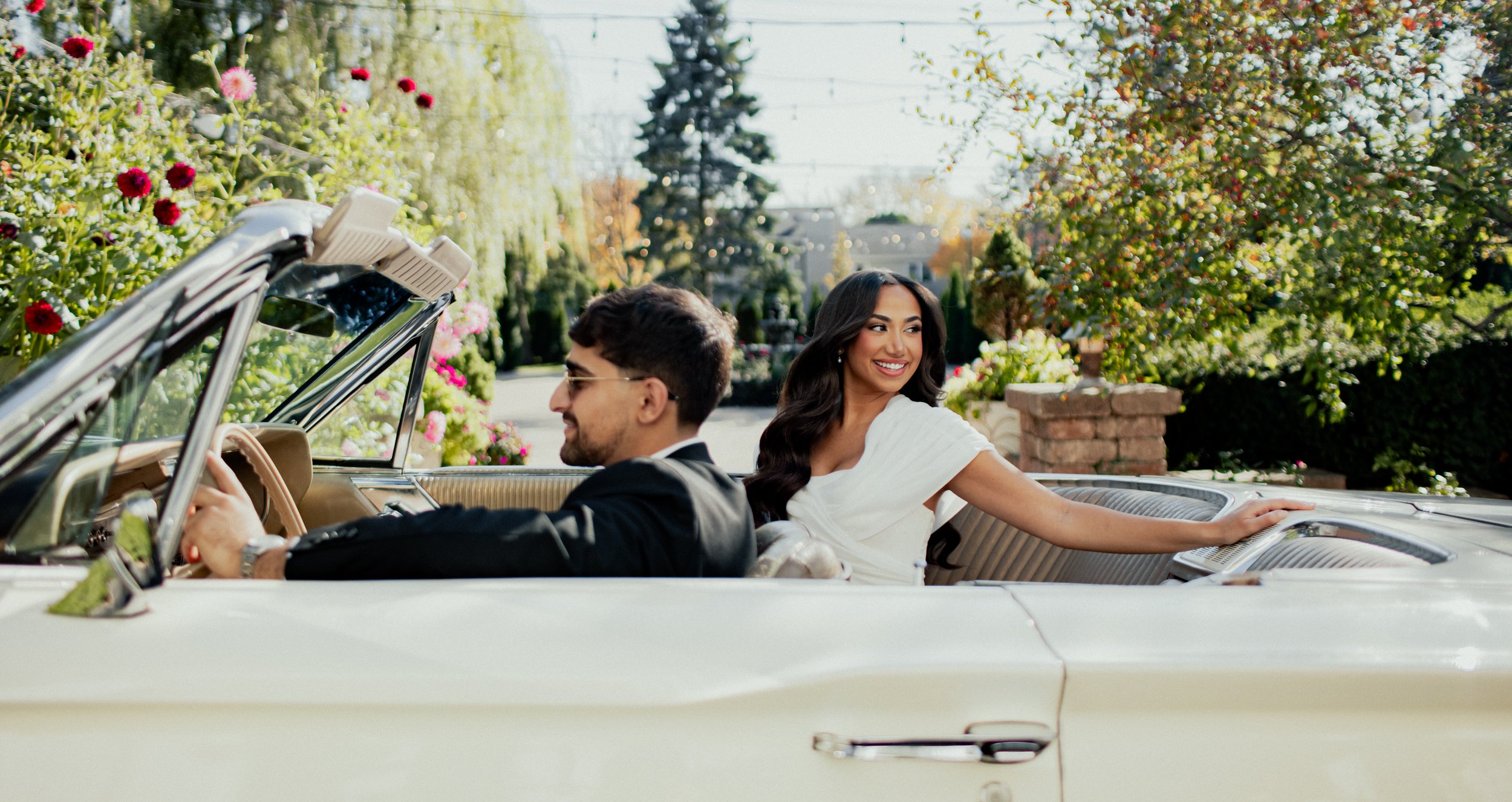 A young man and woman enjoying a ride in a vintage white convertible car, with the woman smiling and holding onto the side of the car. They are outdoors among lush greenery and flowers, with sunlight illuminating the scene.