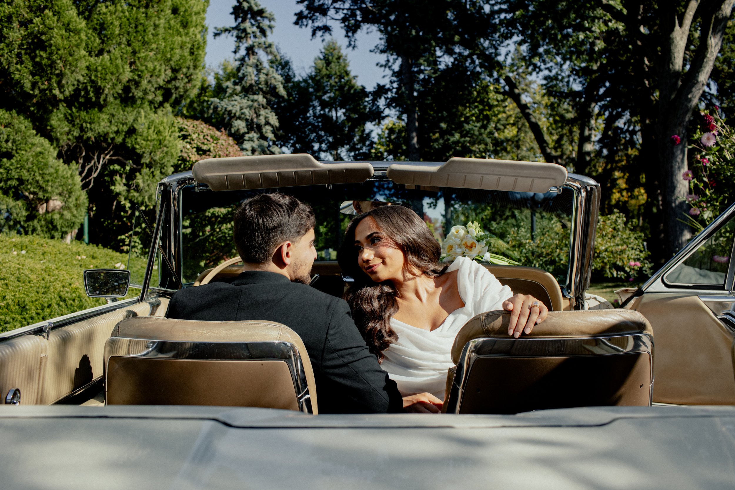 A newlywed couple sitting in the backseat of a vintage convertible car, sharing a romantic moment outdoors with trees and greenery in the background.