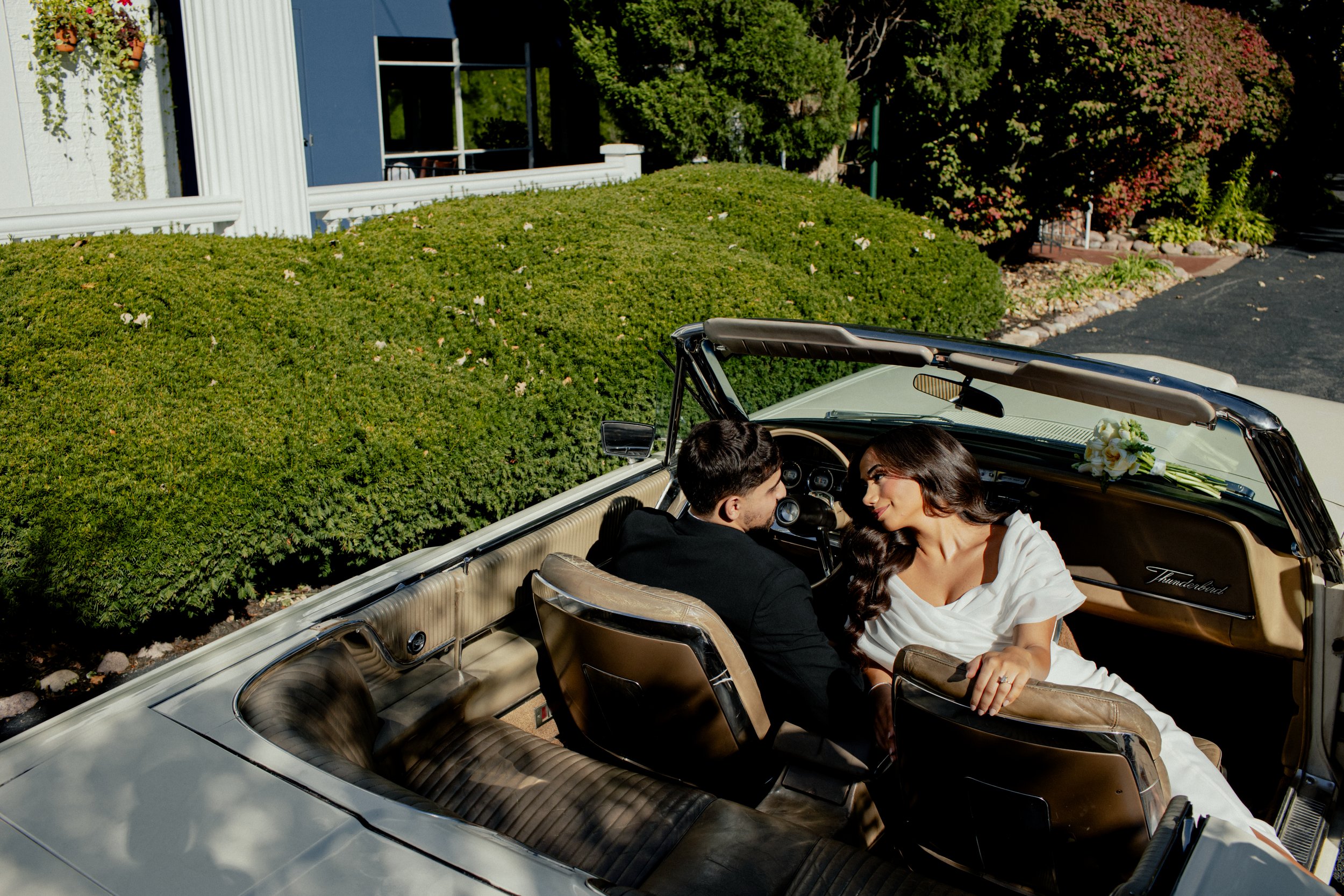 A couple is sitting in a vintage convertible car, looking at each other affectionately. The woman is wearing a white dress and the man is in a dark suit. There is a bouquet of flowers on the dashboard. They are parked near a lush green area with bush