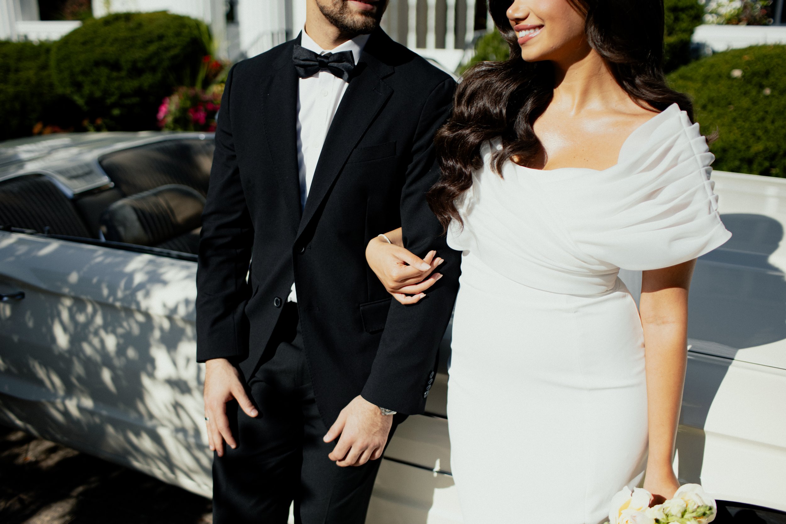 A man and woman dressed in formal attire standing next to a vintage white car. The man is wearing a black tuxedo with a bow tie, and the woman is wearing a white dress with pleated sleeves, holding a bouquet.