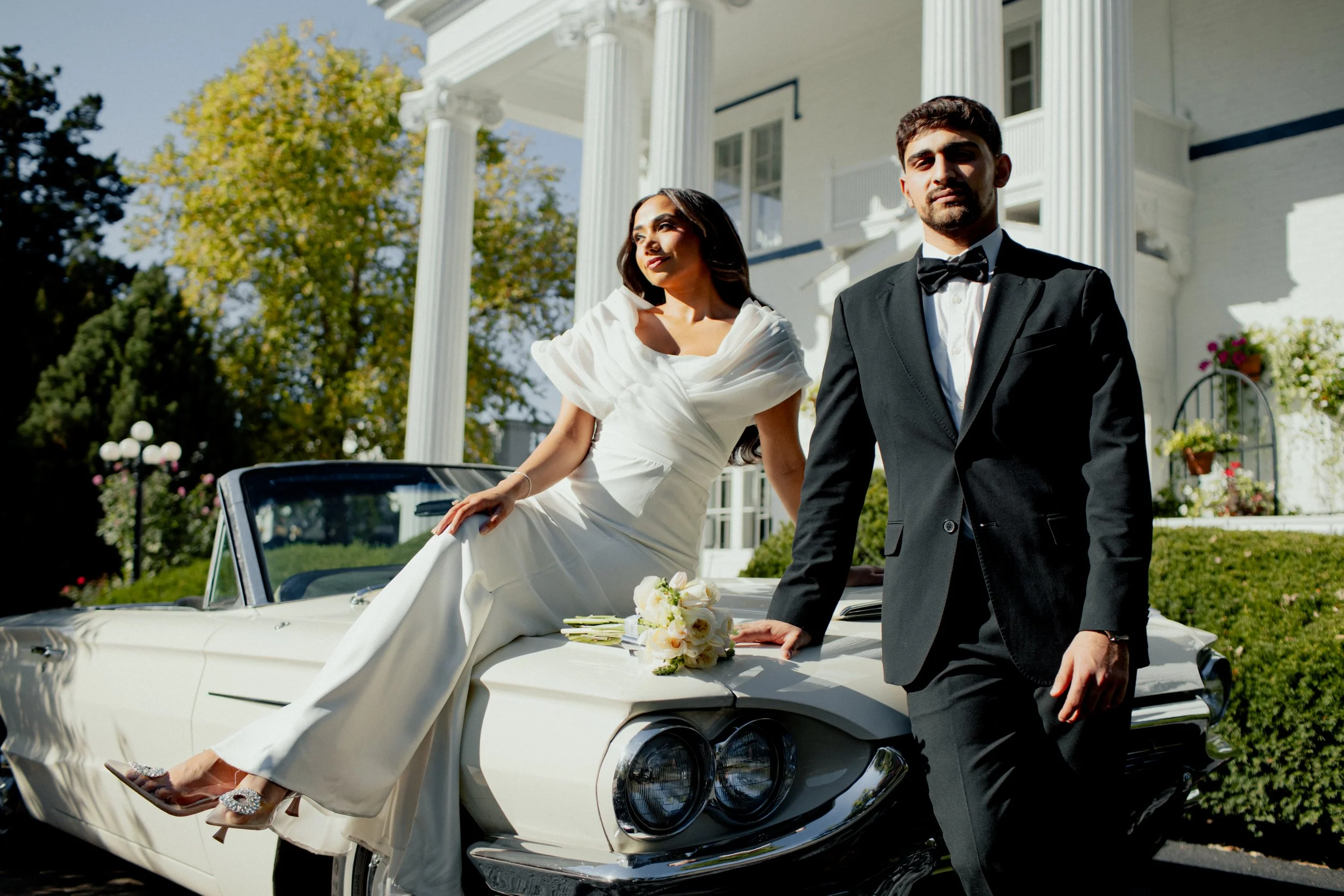 A bride and groom in wedding attire leaning on a vintage white convertible car outside a large white house with columns and green landscaping.