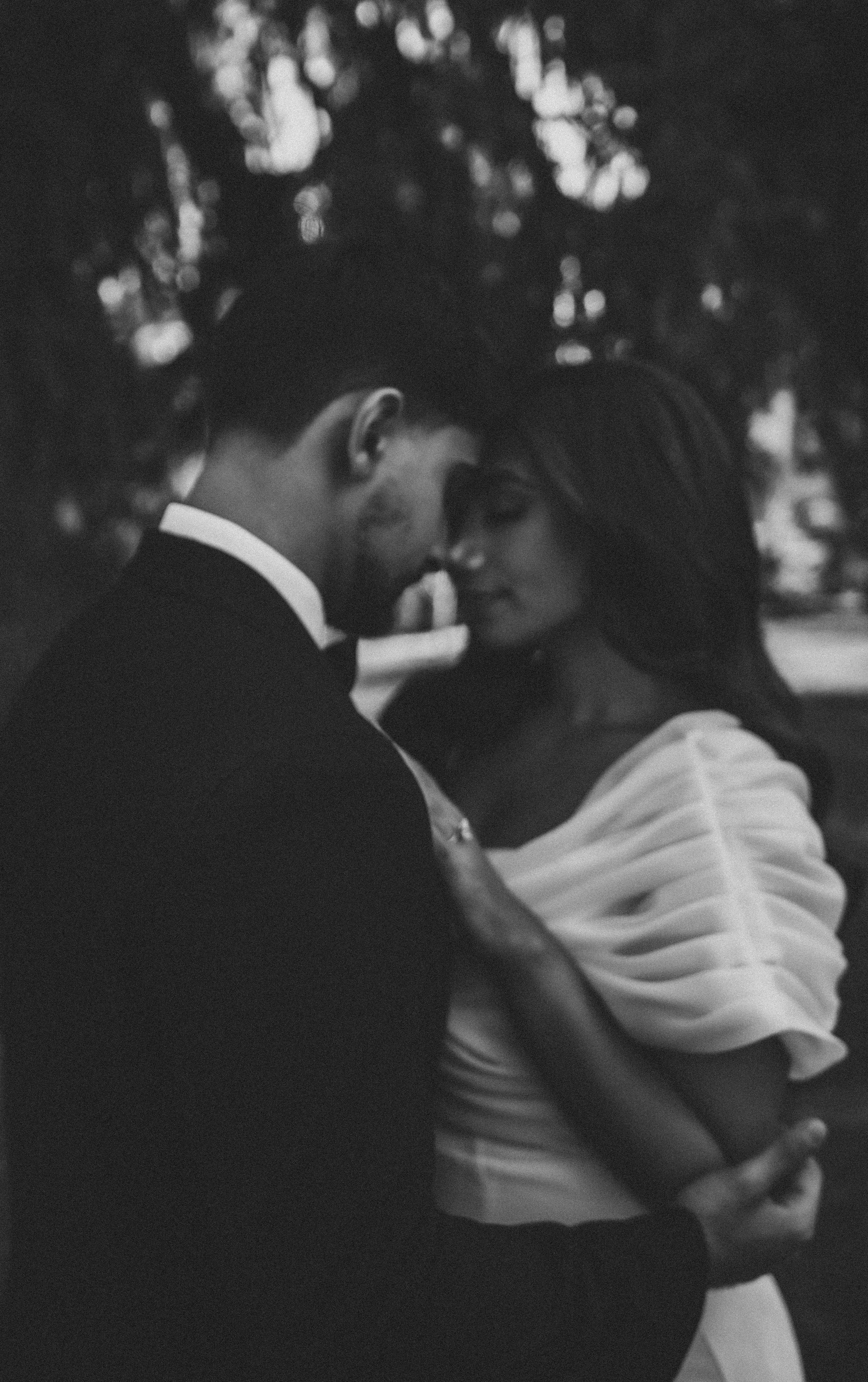 A black and white photo of a couple embracing outdoors, with the man in a suit and the woman in a dress, their foreheads touching under a background of blurred trees.