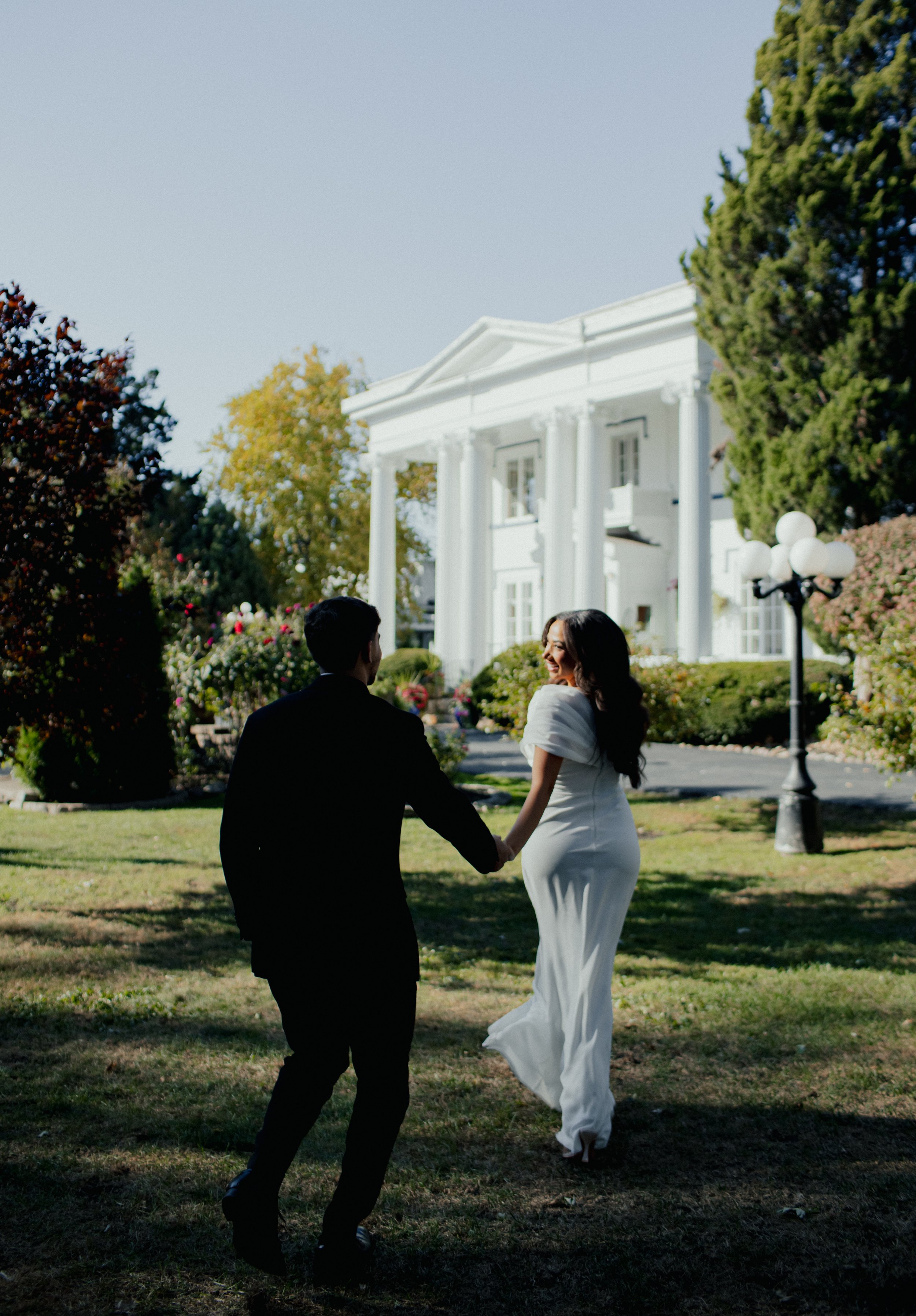 A man and woman holding hands and dancing outside a white mansion on a sunny day, with trees and bushes surrounding them.