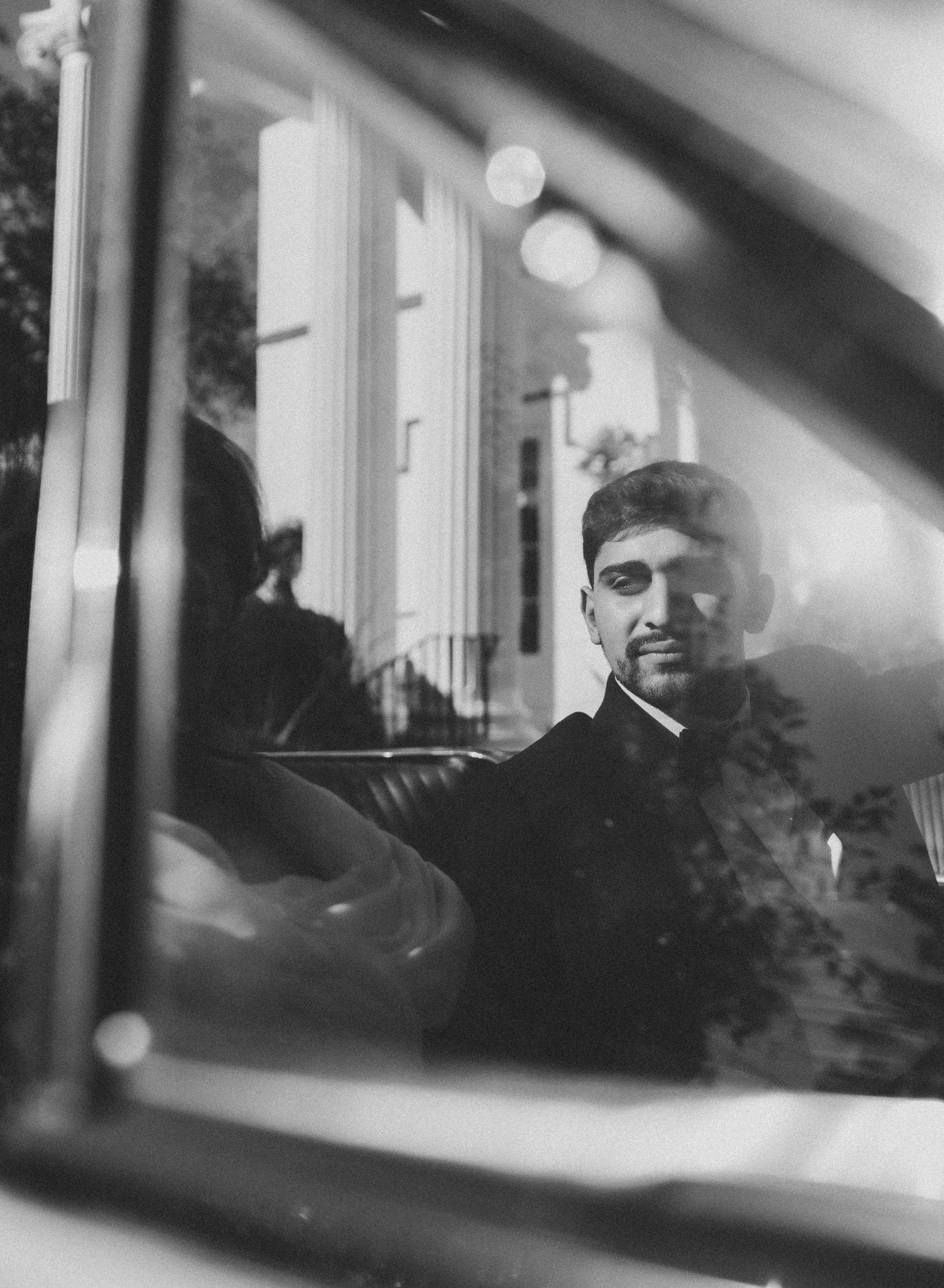 A black-and-white photo of a young man wearing a tuxedo, sitting inside a vintage car, looking through the window. The background shows a building with large columns and trees, reflected in the window.