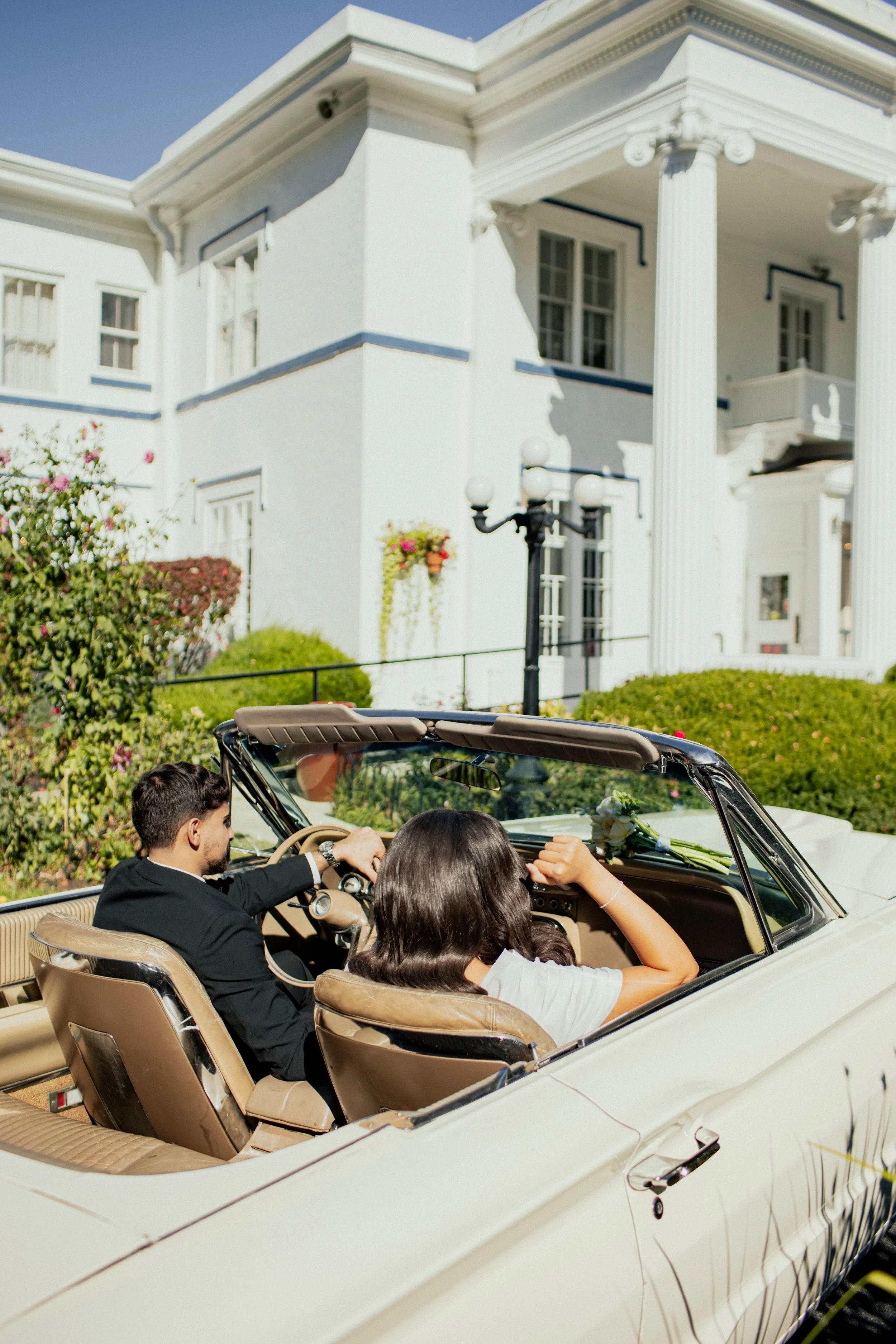 A man and a woman sitting in a vintage convertible car parked in front of a large white house with columns, greenery, and flowers.