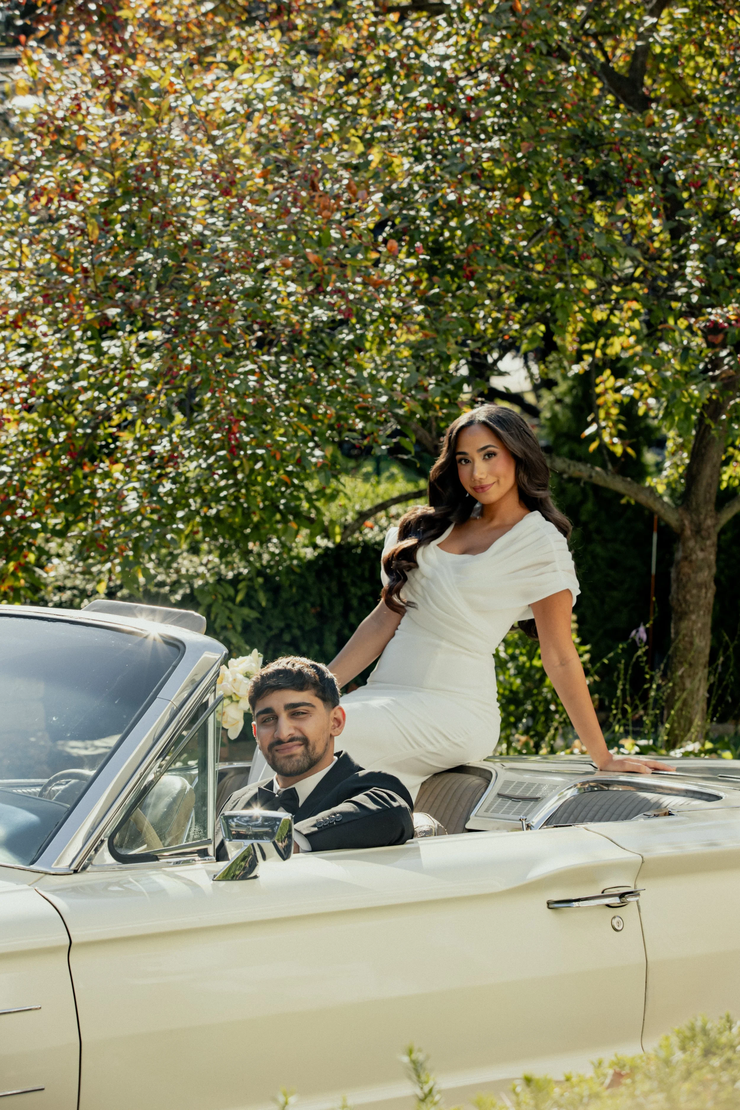 A young couple dressed in formal attire sitting in a vintage convertible car outdoors surrounded by greenery, with the woman sitting on the back of the car and the man in the driver’s seat.