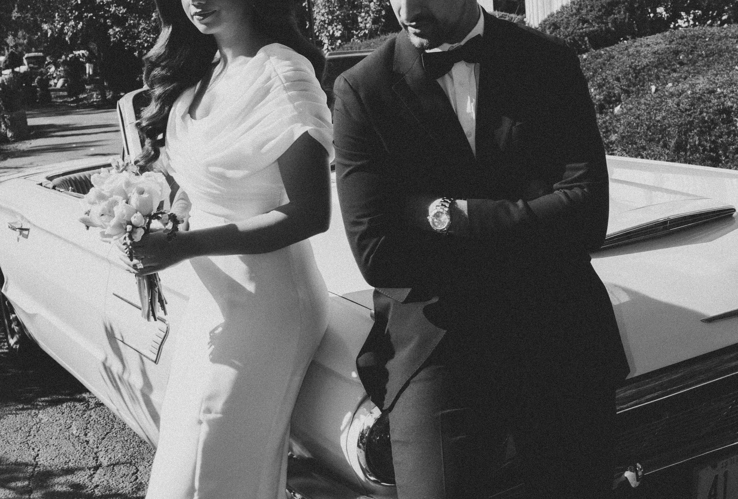 A couple dressed in formal wedding attire, standing beside a classic white convertible car. The woman is holding a bouquet of flowers, and the man has his arms crossed, wearing a tuxedo with a bow tie and a watch.