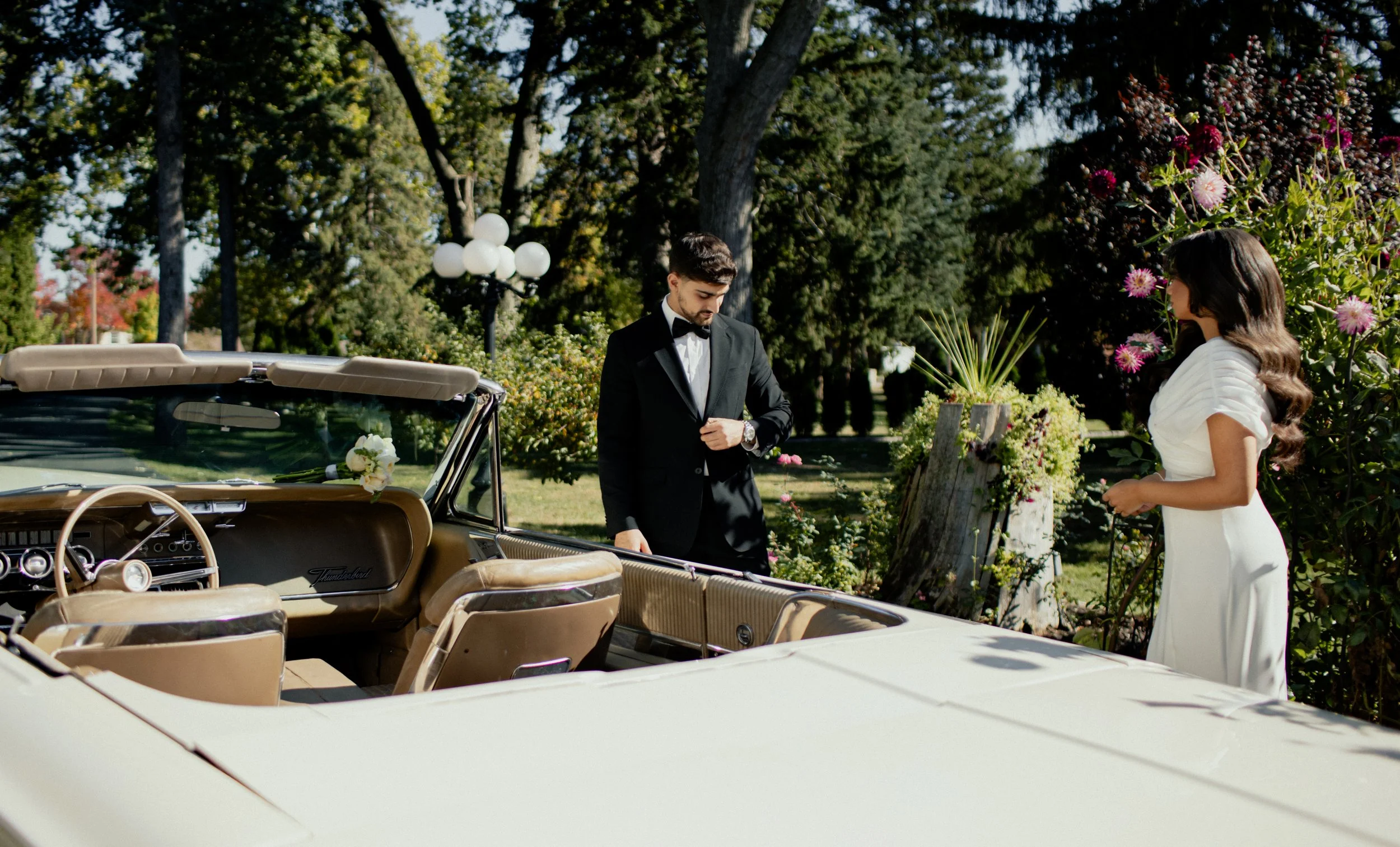 A man and woman dressed in formal wedding attire standing next to a vintage convertible car in a lush garden with trees and flowers.