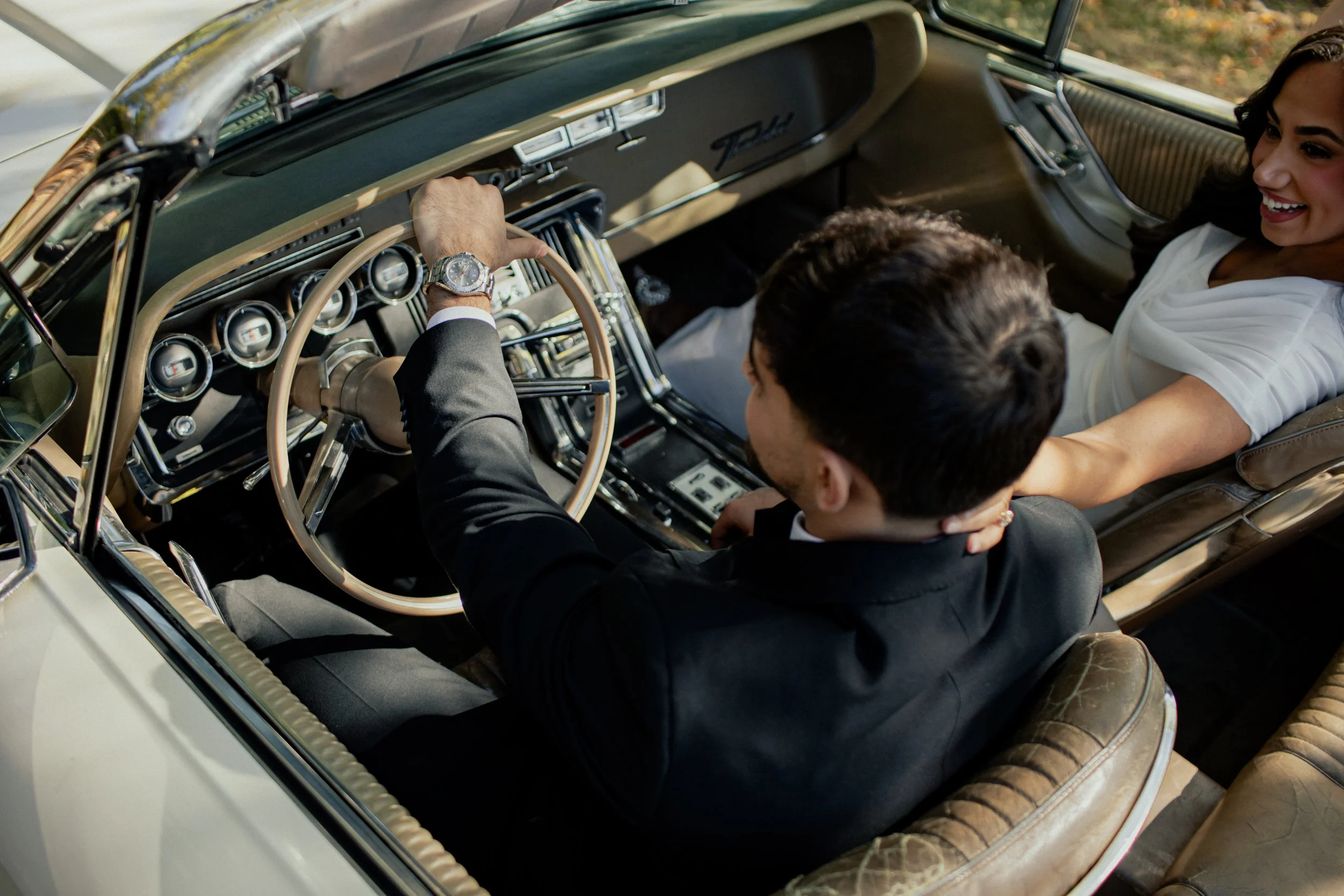 A man and woman sitting in a vintage car, with the man in the driver's seat and the woman sitting beside him, smiling and reaching out to his face.