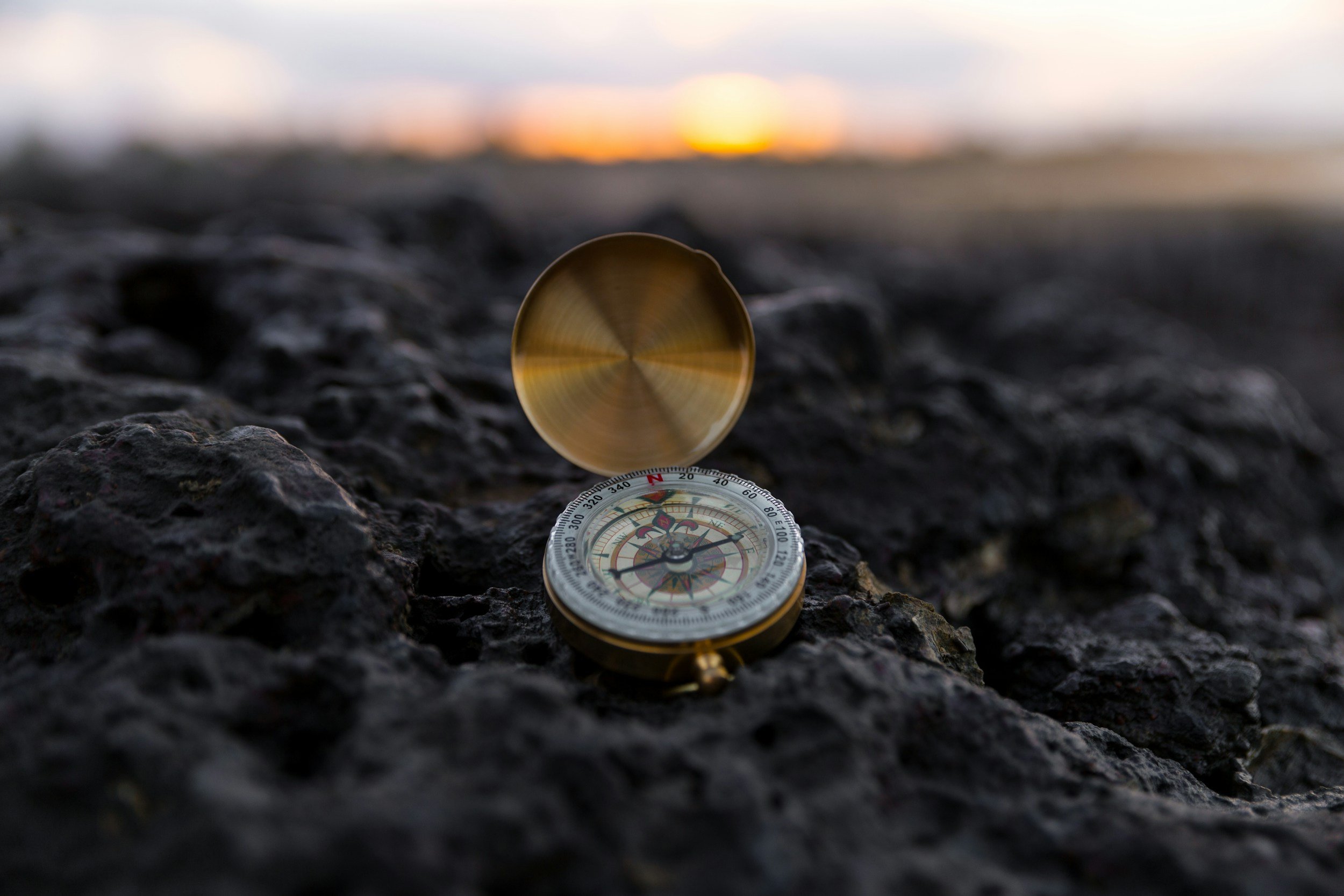 A compass lying on dark, rough rocks with a sunset or sunrise in the blurred background.