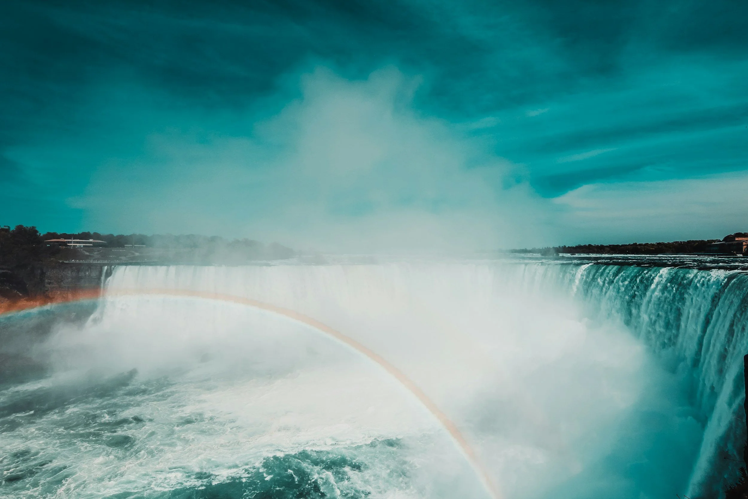 View of Niagara Falls with mist rising from the cascading water, overcast sky, and some buildings visible on the distant shoreline.