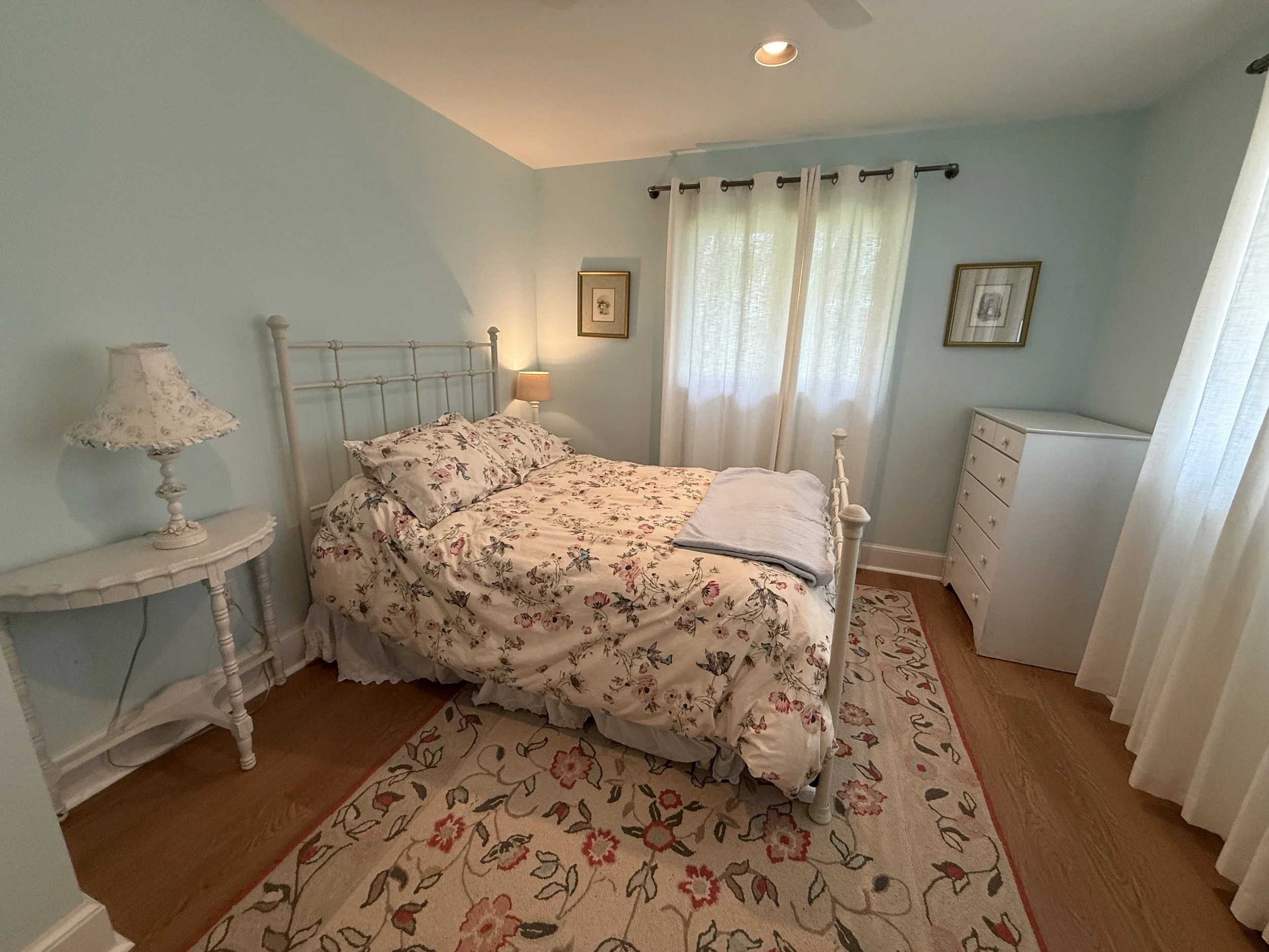 A cozy bedroom with a white metal bed frame, floral bedding, a matching bedside lamp, and curtains over a window. The room features framed artwork and a patterned area rug on wooden flooring.