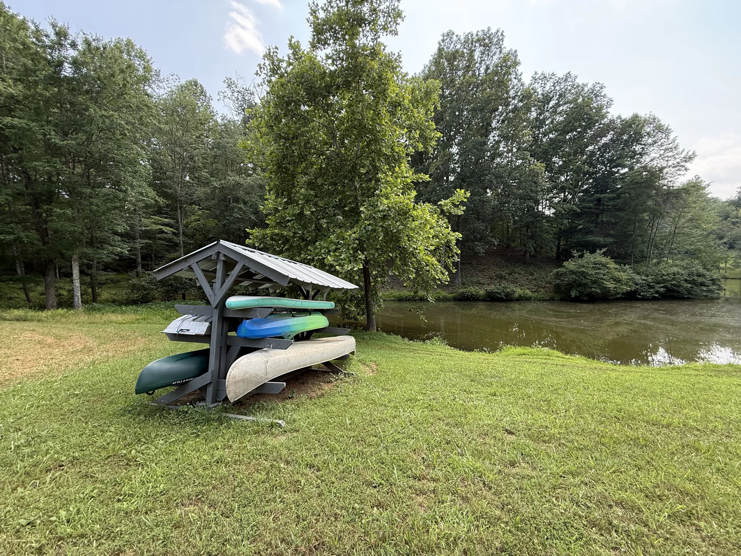 A lakeside scene with a small boat dock and a covered boat storage area, surrounded by trees and grass, with the lake and sky in the background.