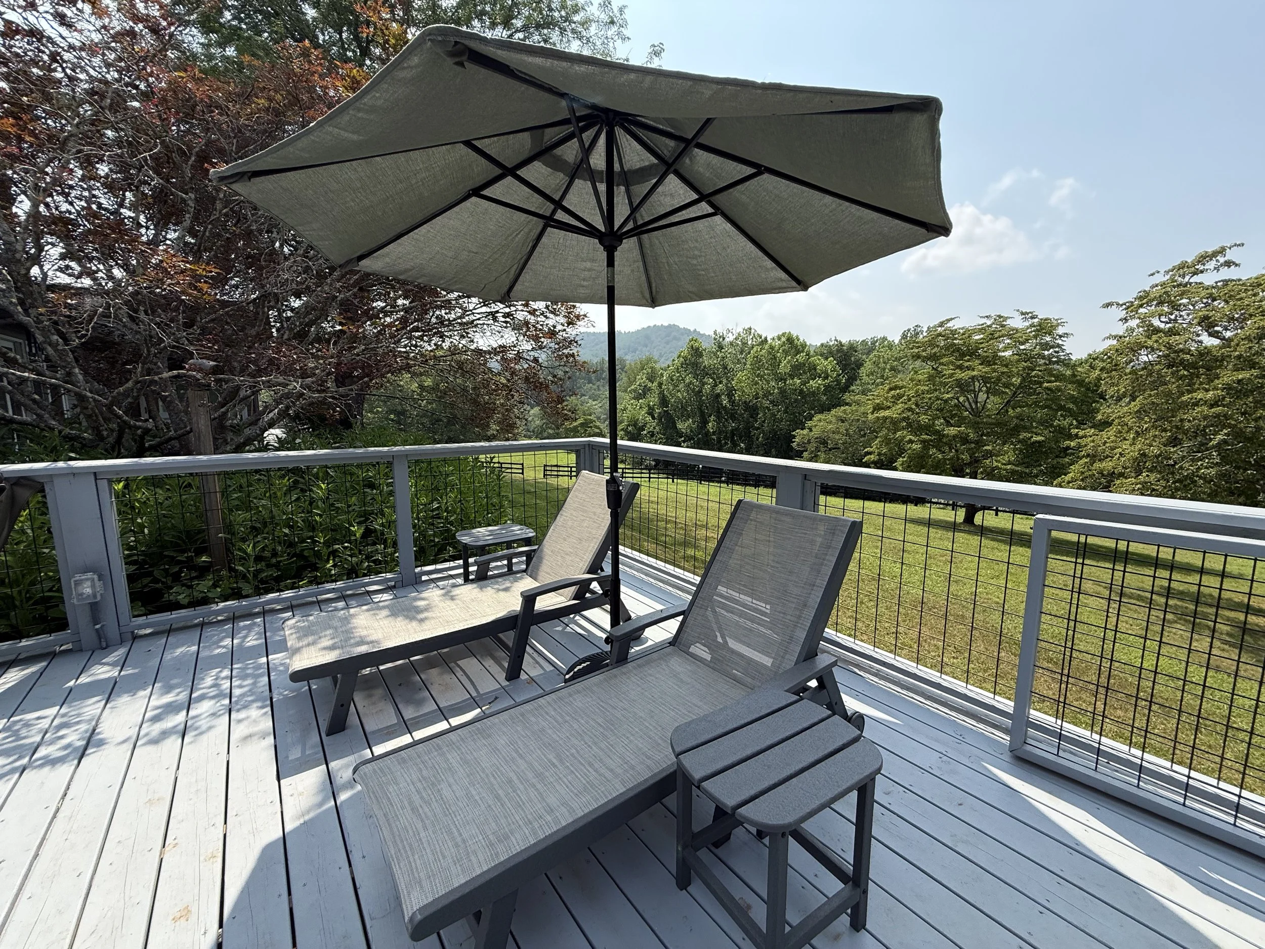 A wooden deck with patio furniture and umbrellas, surrounded by trees and a view of distant mountains under a clear blue sky.