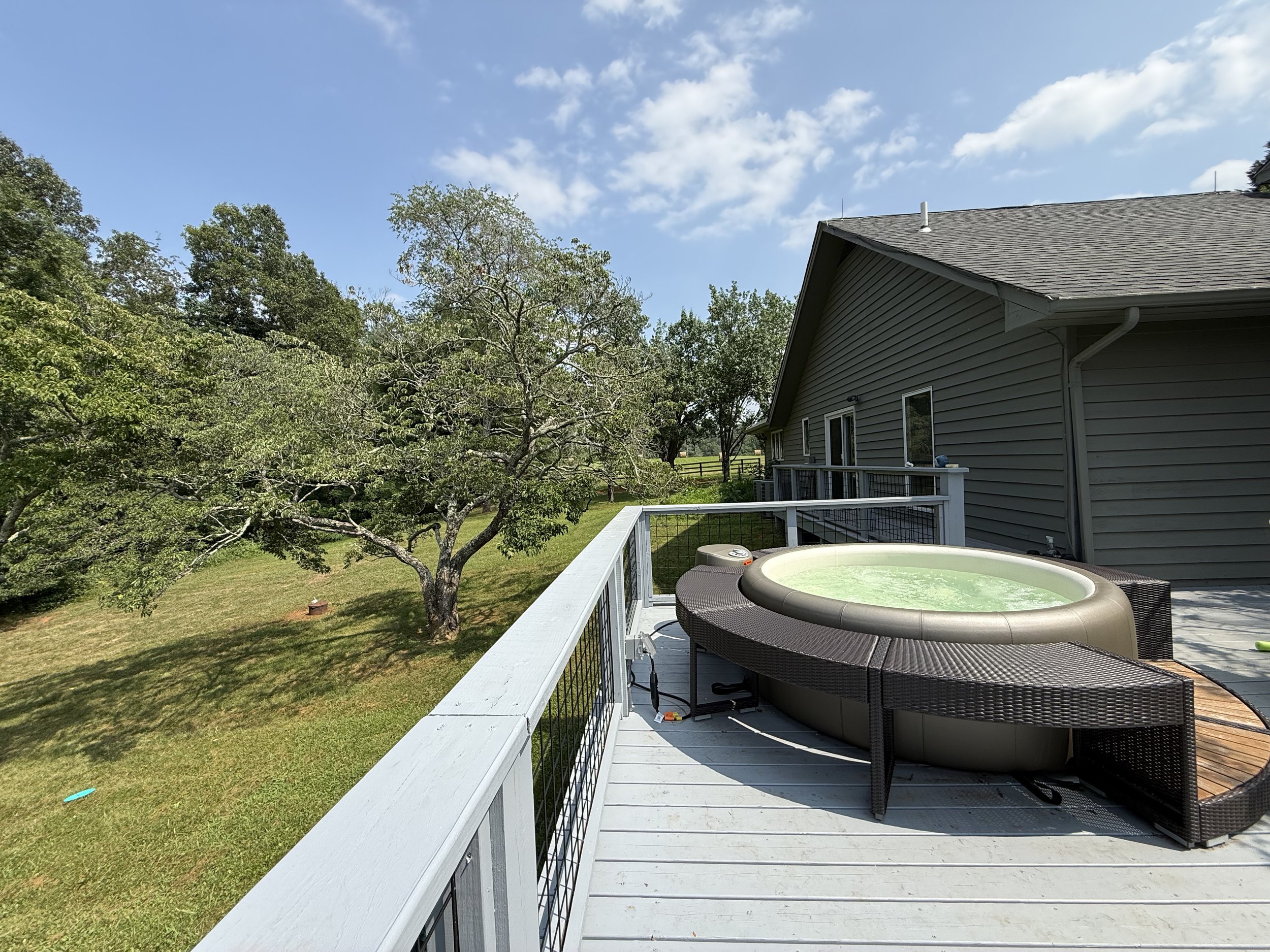 Backyard patio with hot tub, wicker chairs, and a view of green lawn, trees, and a house with gray siding and a shaded roof on a sunny day.