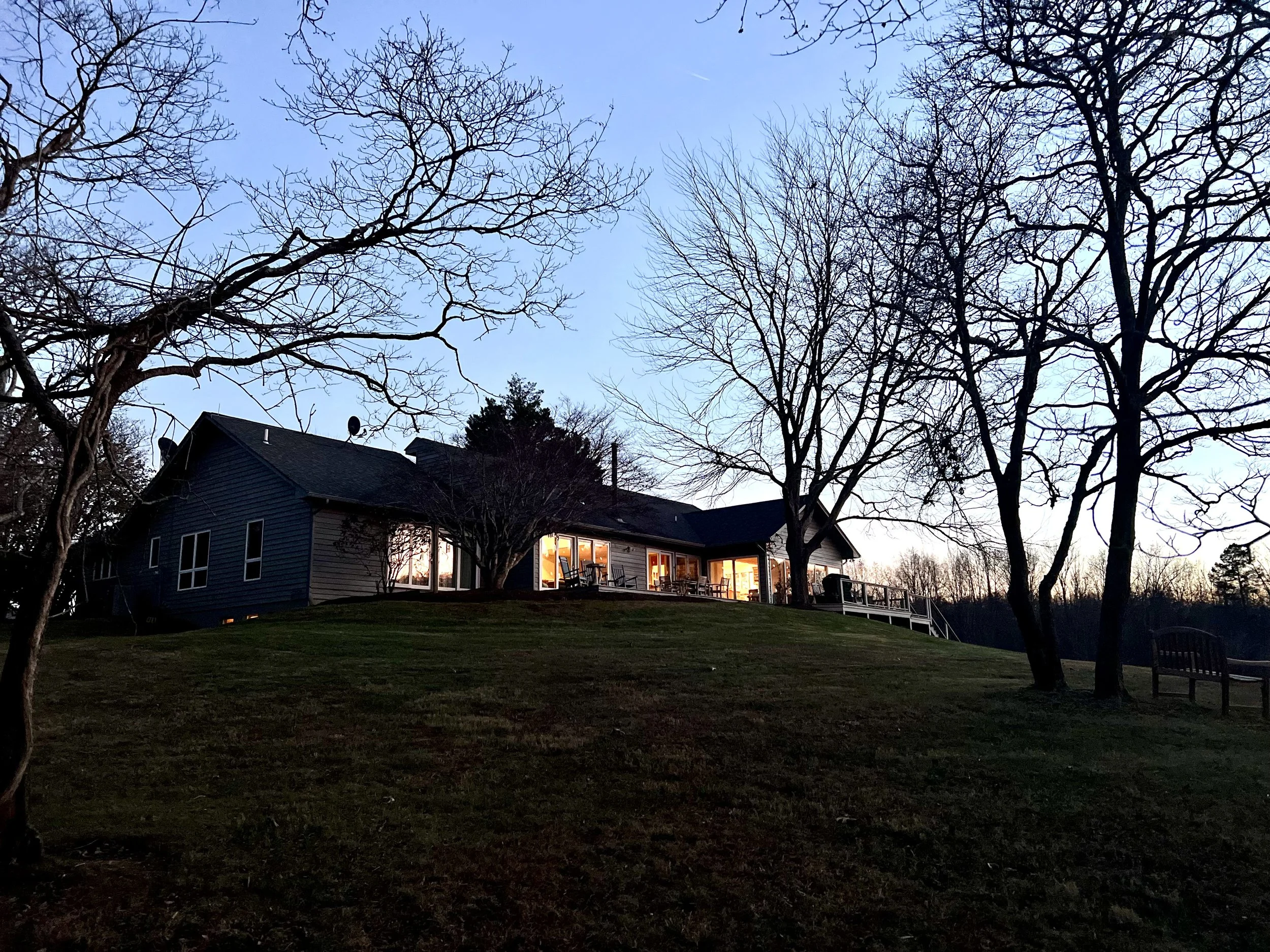 Retreat property in the Shenandoah Mountains. A house with illuminated windows surrounded by leafless trees in the evening or early morning sky.