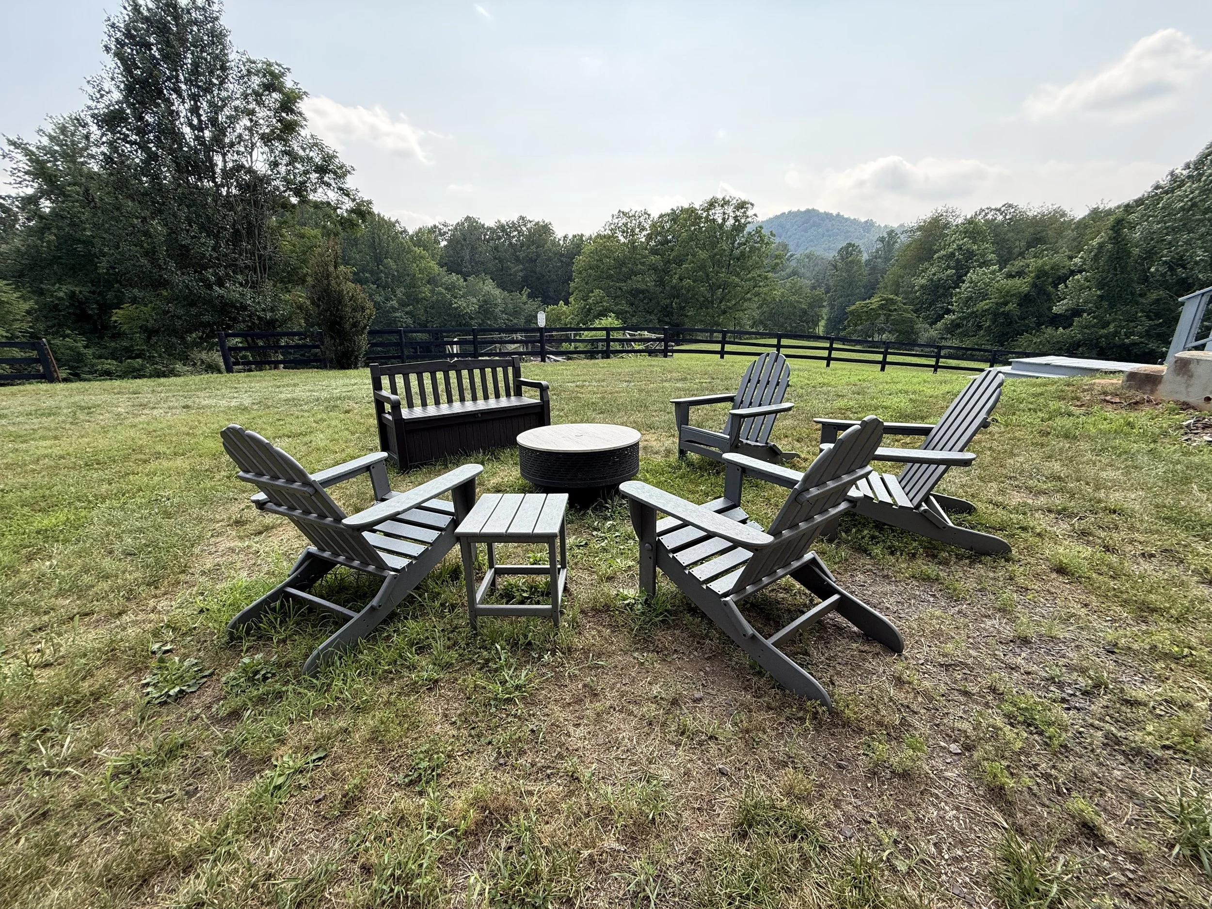 Outdoor seating area with Adirondack chairs surrounding a fire pit on a grassy lawn, with a backdrop of trees and distant mountains under a partly cloudy sky.