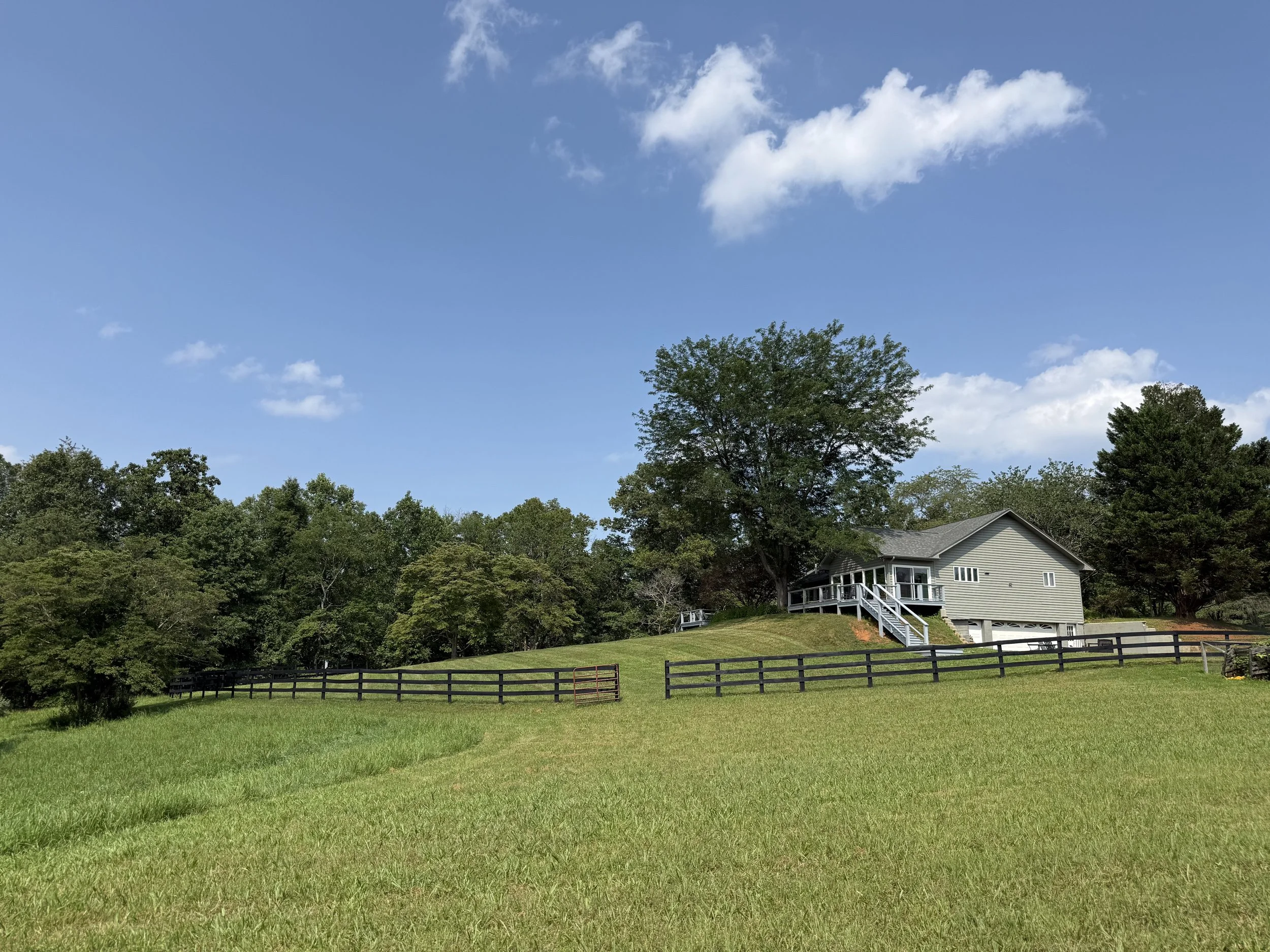 A two-story house with a porch and stairs, surrounded by green trees and a grassy yard, under a blue sky with some clouds.