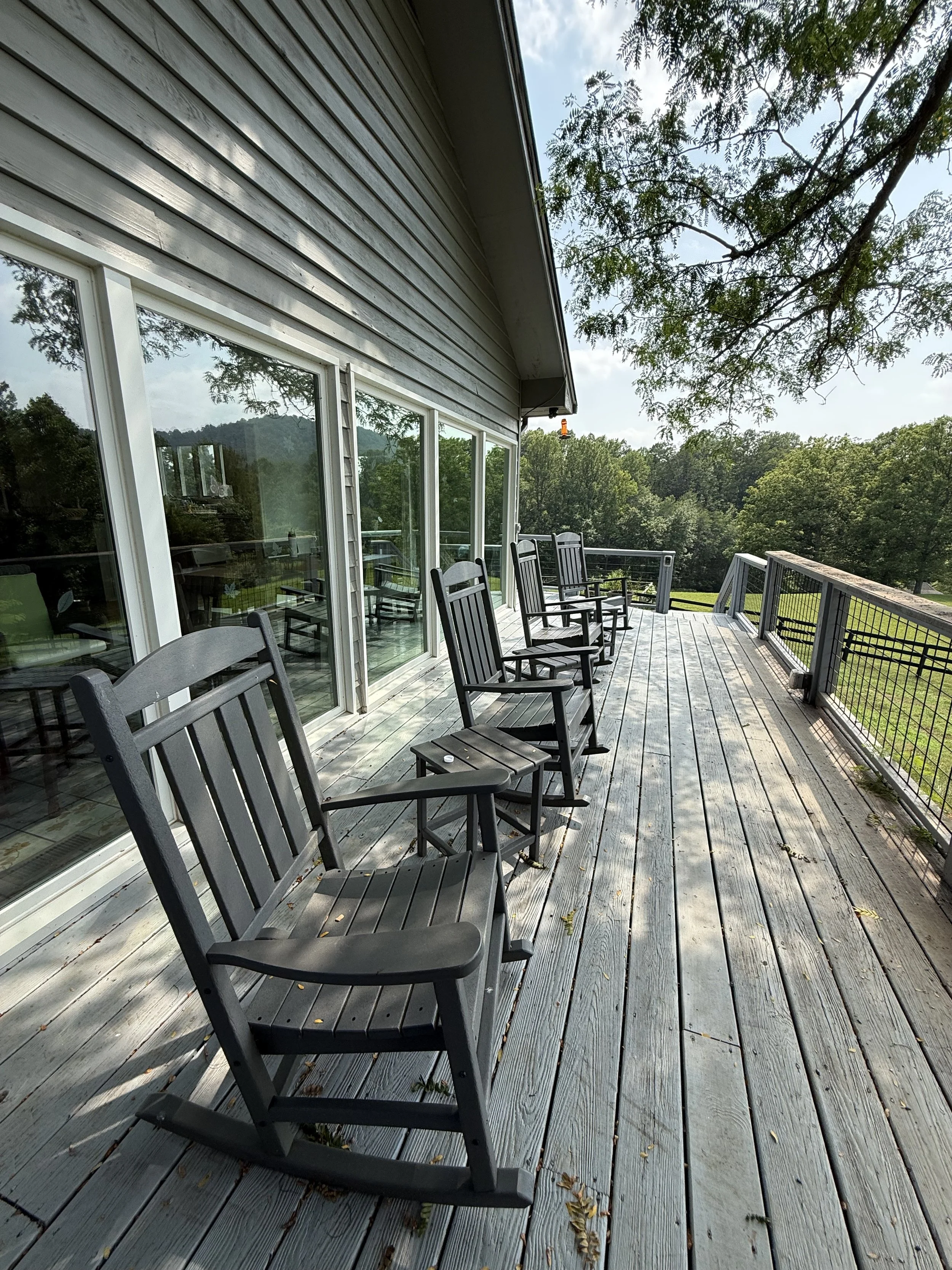 An outdoor wooden deck with black chairs, a railing, and glass sliding doors leading into a house, surrounded by trees and a blue sky with clouds.