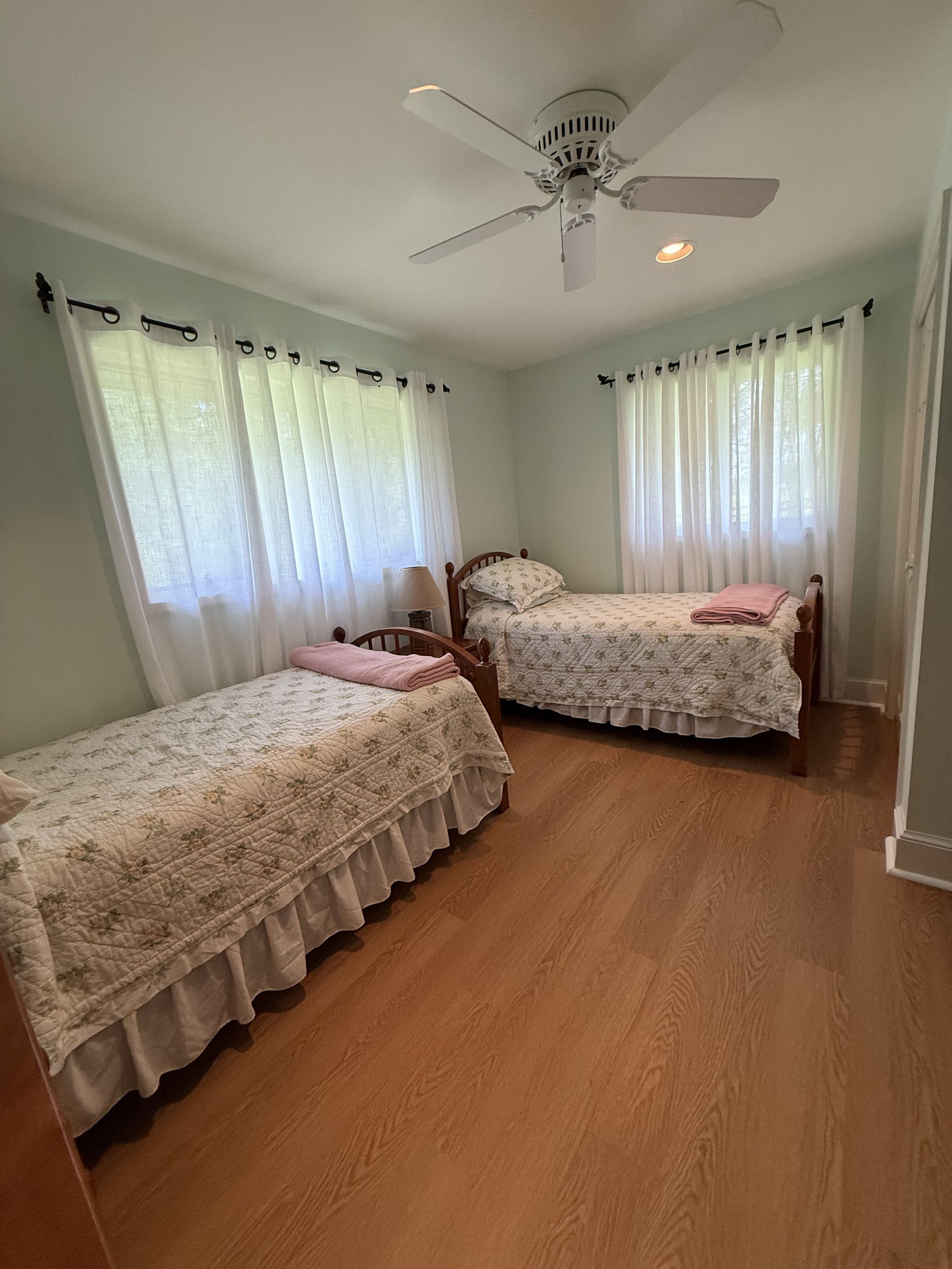 A bedroom with two twin beds with floral quilts, pink towels, and pillows, green walls, white curtains, and a ceiling fan.