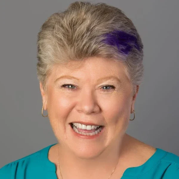 A smiling middle-aged woman with short gray hair and a purple streak, wearing a turquoise top and hoop earrings, against a gray background.