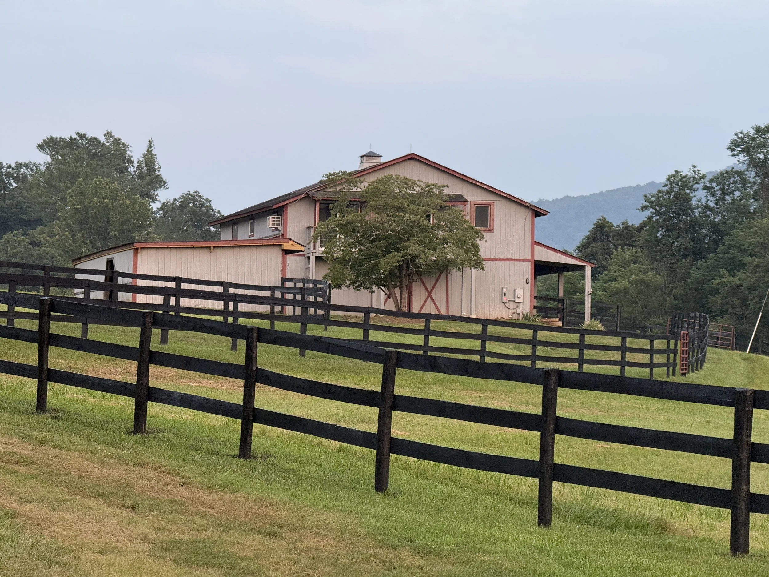 A farm scene with a wooden barn, surrounded by black fencing, green grass, and trees, with mountains in the background and a cloudy sky.