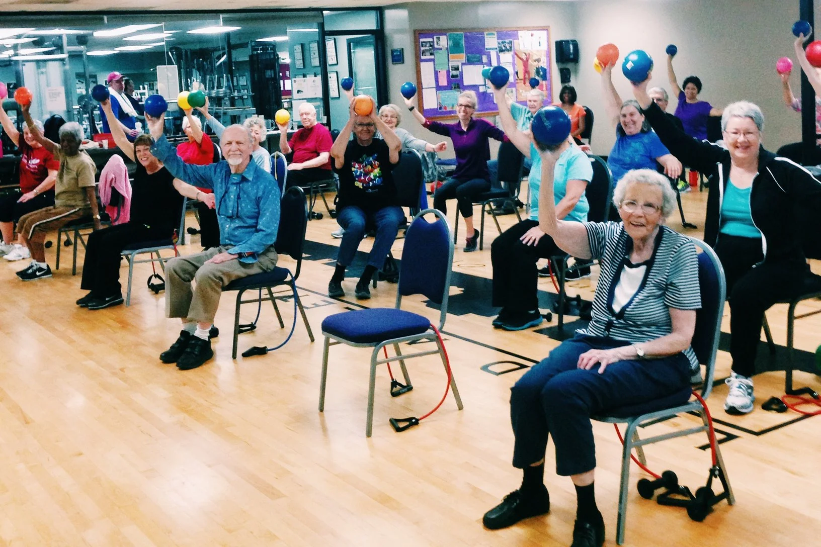Group of seniors participating in a seated exercise class, holding colorful weights and smiling, in a gym setting.