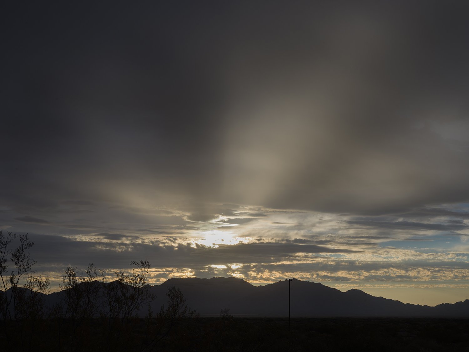 Dark cloudy sky over a mountain range with silhouette of sparse trees and a single utility pole, during sunset or sunrise.