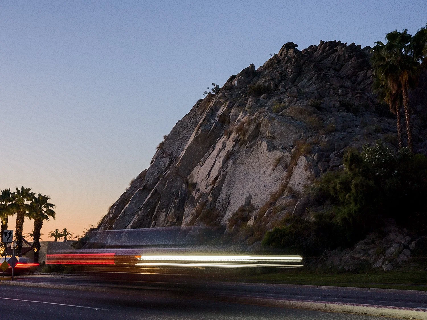 The image shows a large rocky hillside with palm trees near the road at sunset, with light trails from moving vehicles in the foreground.