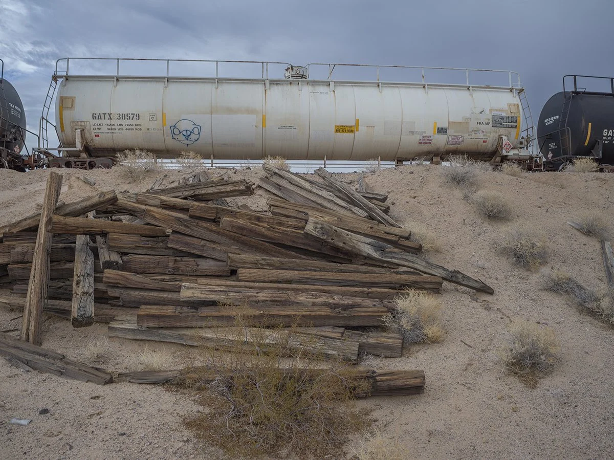 A white railroad tanker car on tracks above a pile of weathered wooden planks in a desert landscape with small bushes and a cloudy sky.