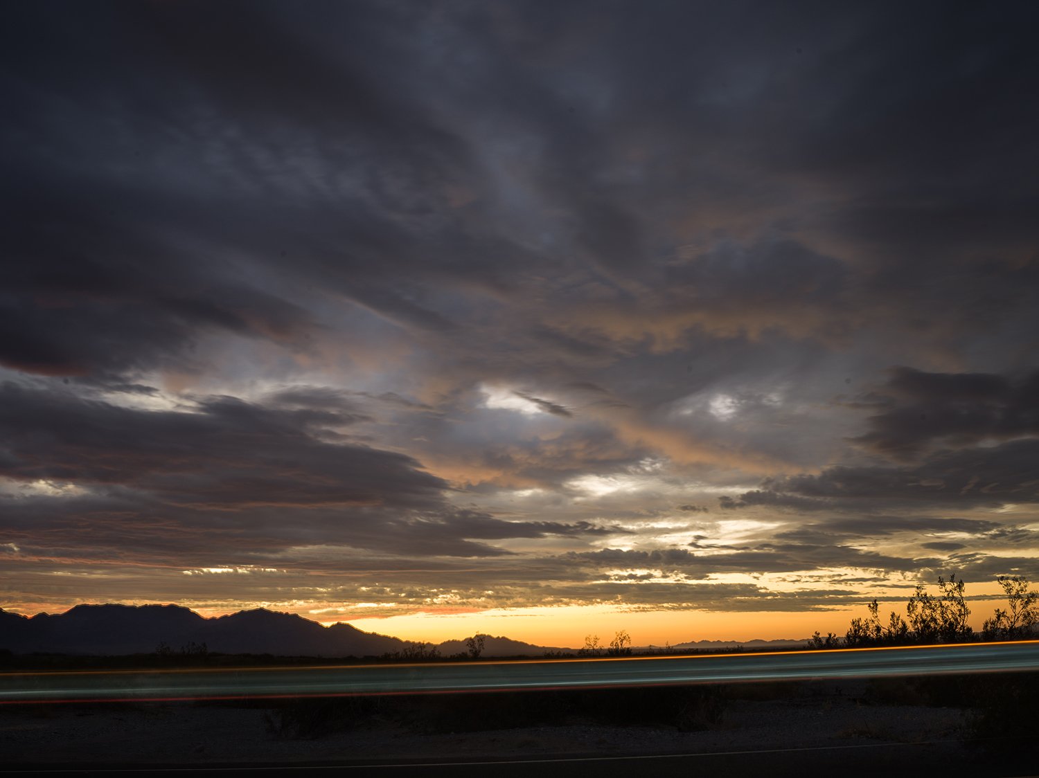 A dramatic sunset over a mountain range with dark clouds overhead and light trails from vehicles on a highway in the foreground.